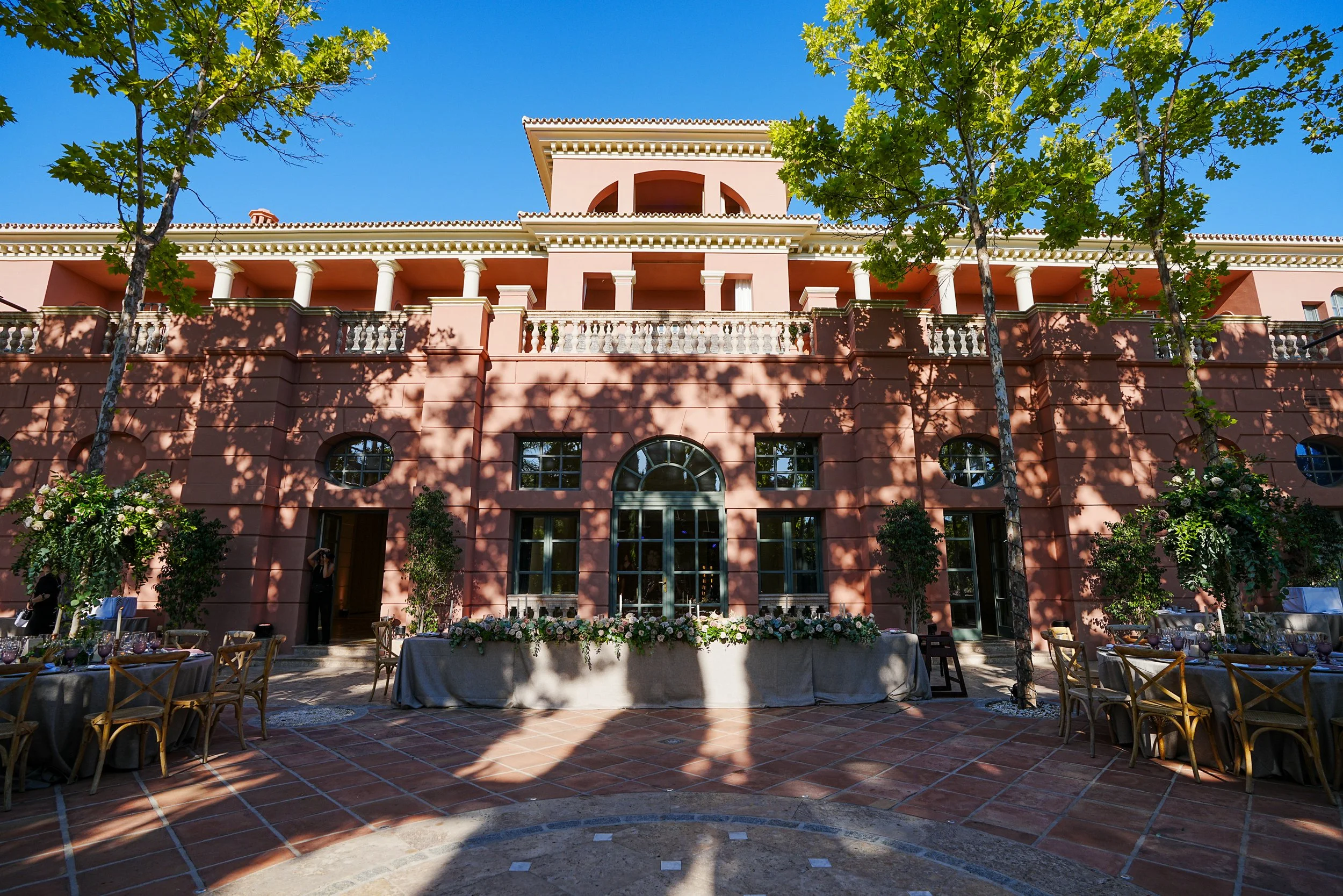 An elegant terracotta-colored building with arches, balconies with balustrades, and a central tower under a bright blue sky, surrounded by green trees.