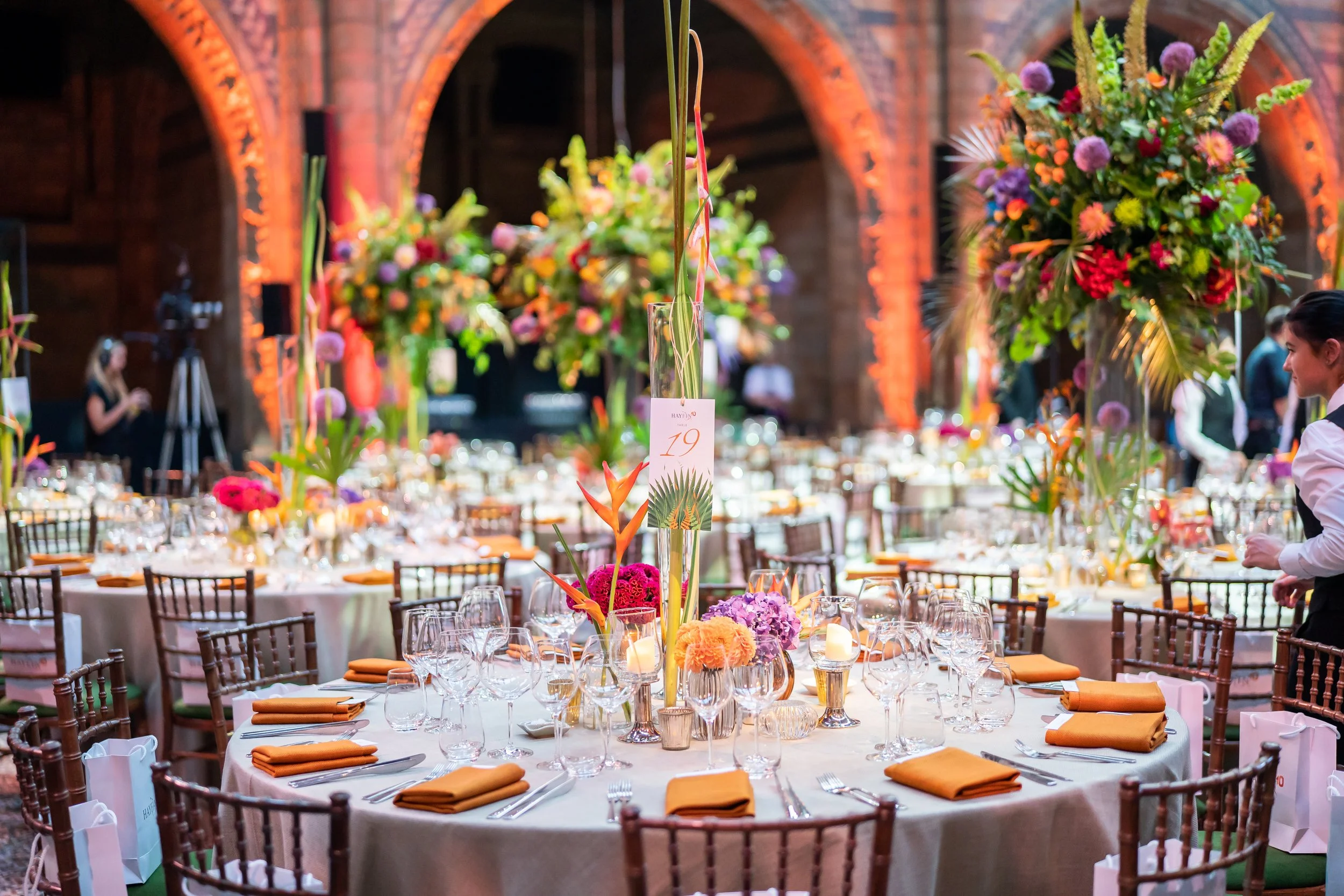A banquet hall decorated with colorful floral centerpieces on elegantly set tables, with guests and waitstaff visible in the background, and exposed brick arches in the architecture at The Natural History Museum