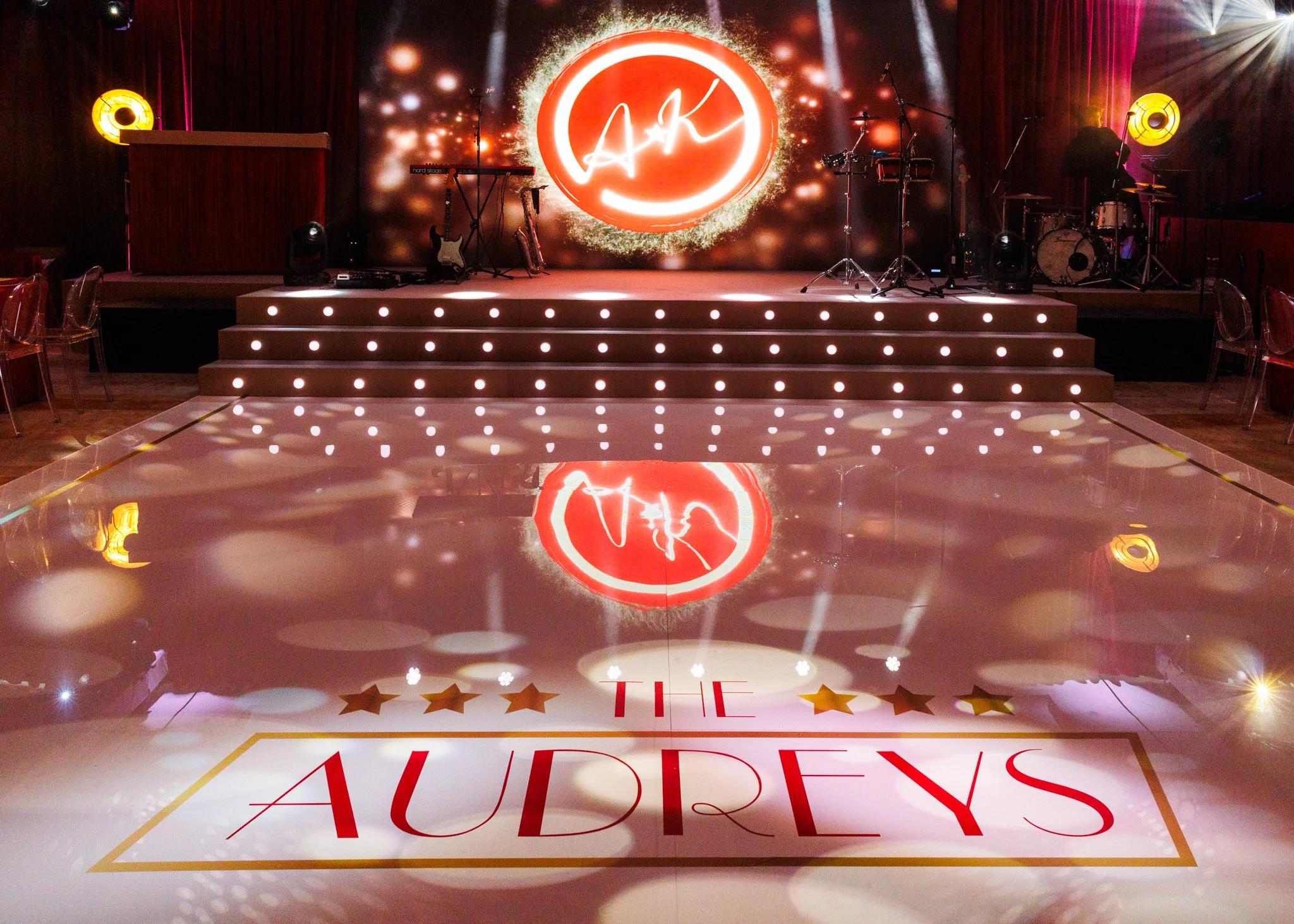 Stage with musical instruments and a large illuminated red circle with initials 'AK'. The stage is decorated with bright lights, and there is a reflection of the scene on the shiny floor. The words 'THE' and 'AWARDS' are partially visible on the floo