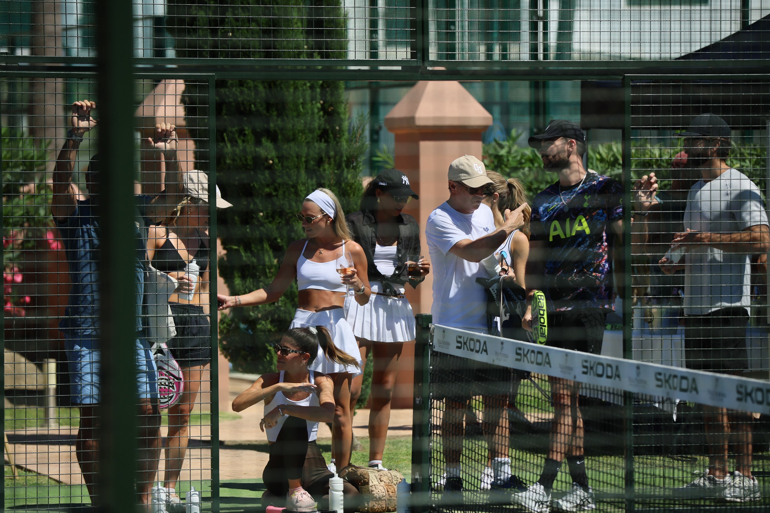 People gathered at a tennis court, standing next to a net with a Skoda banner, some holding drinks, amid greenery and a building in the background.