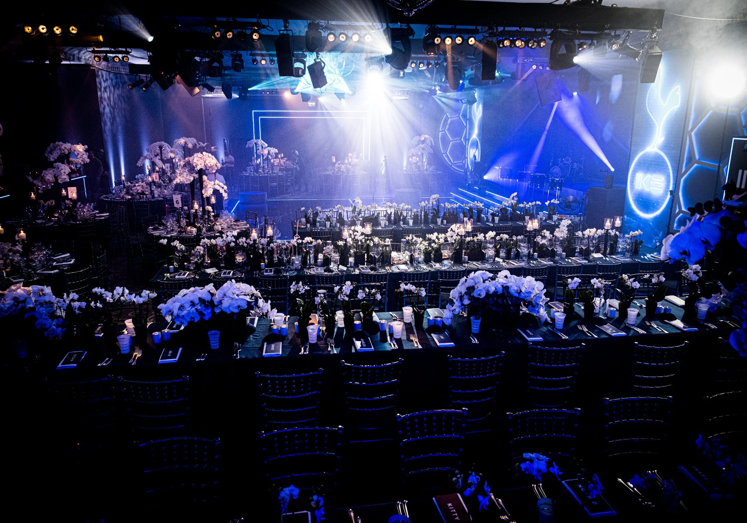 A large event space set up for a formal dinner with many tables decorated with white floral centerpieces and candles, a stage with bright lights, and modern geometric wall decor.