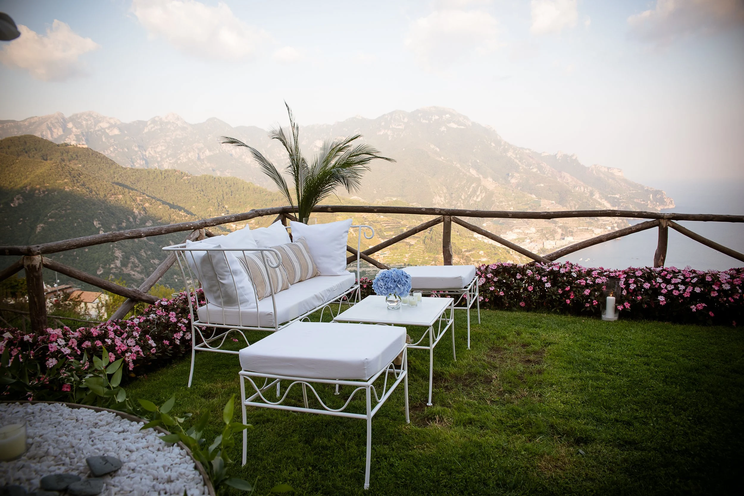 A scenic outdoor terrace with white metal furniture, pillows, and flowers, overlooking mountainous terrain under a cloudy sky.