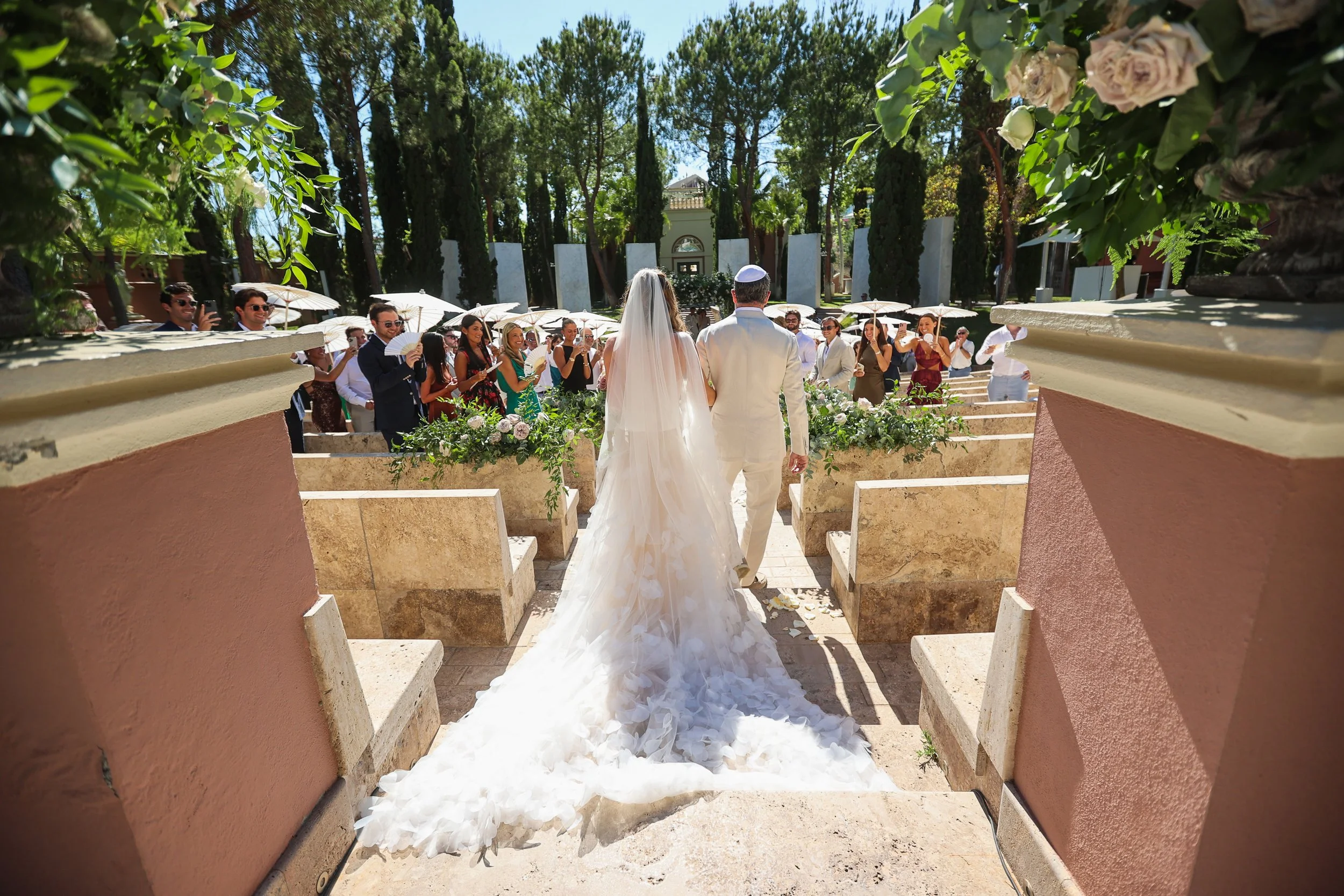 A wedding ceremony outdoors with a bride and groom facing an audience of guests. The bride is in a long white gown with a train and veil, and the groom is in a white suit. Guests are seated under umbrellas, watching the couple. The setting is surroun
