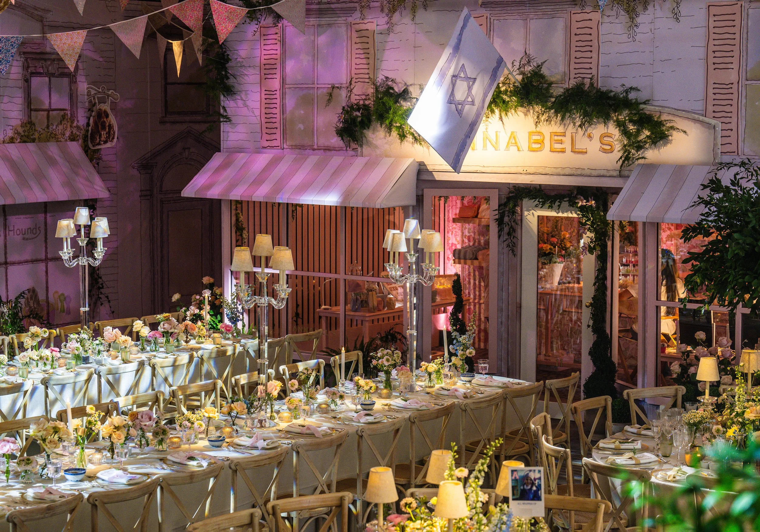A decorated event space with tables set for a Bat Mitzvah, lush floral arrangements, lights, and a wall adorned with a Isreali flag and greenery at Grosvenor House on Park Lane 