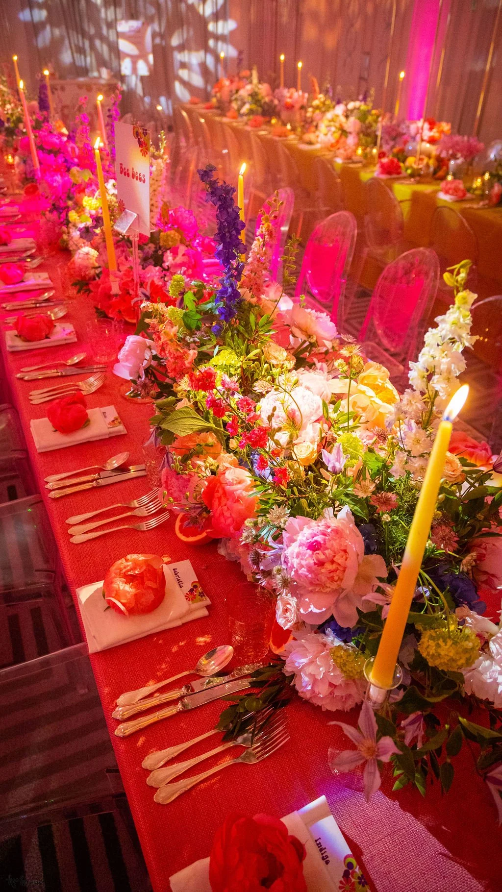 Long banquet table decorated in a 70s vibrant with pink, purple, and white flowers, lit yellow candles, and place settings with silverware, napkins, and pomegranate-shaped objects, in a warmly lit