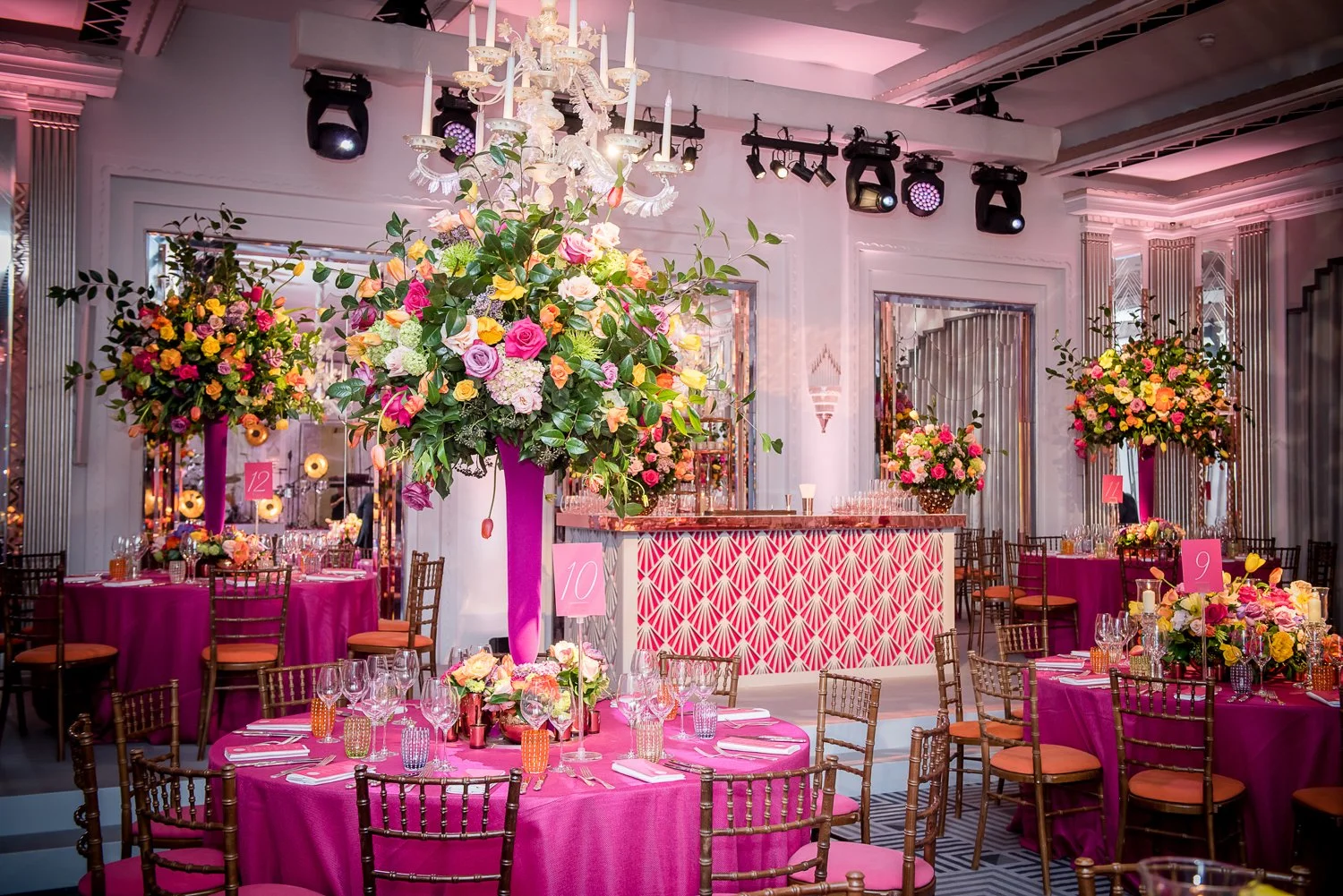 Elegant event space decorated with large floral centerpieces featuring pink, yellow, and white flowers on pink tablecloths, set with glassware and cutlery, and accented by pink table numbers. A chandelier hangs from the ceiling, and the room has a pi