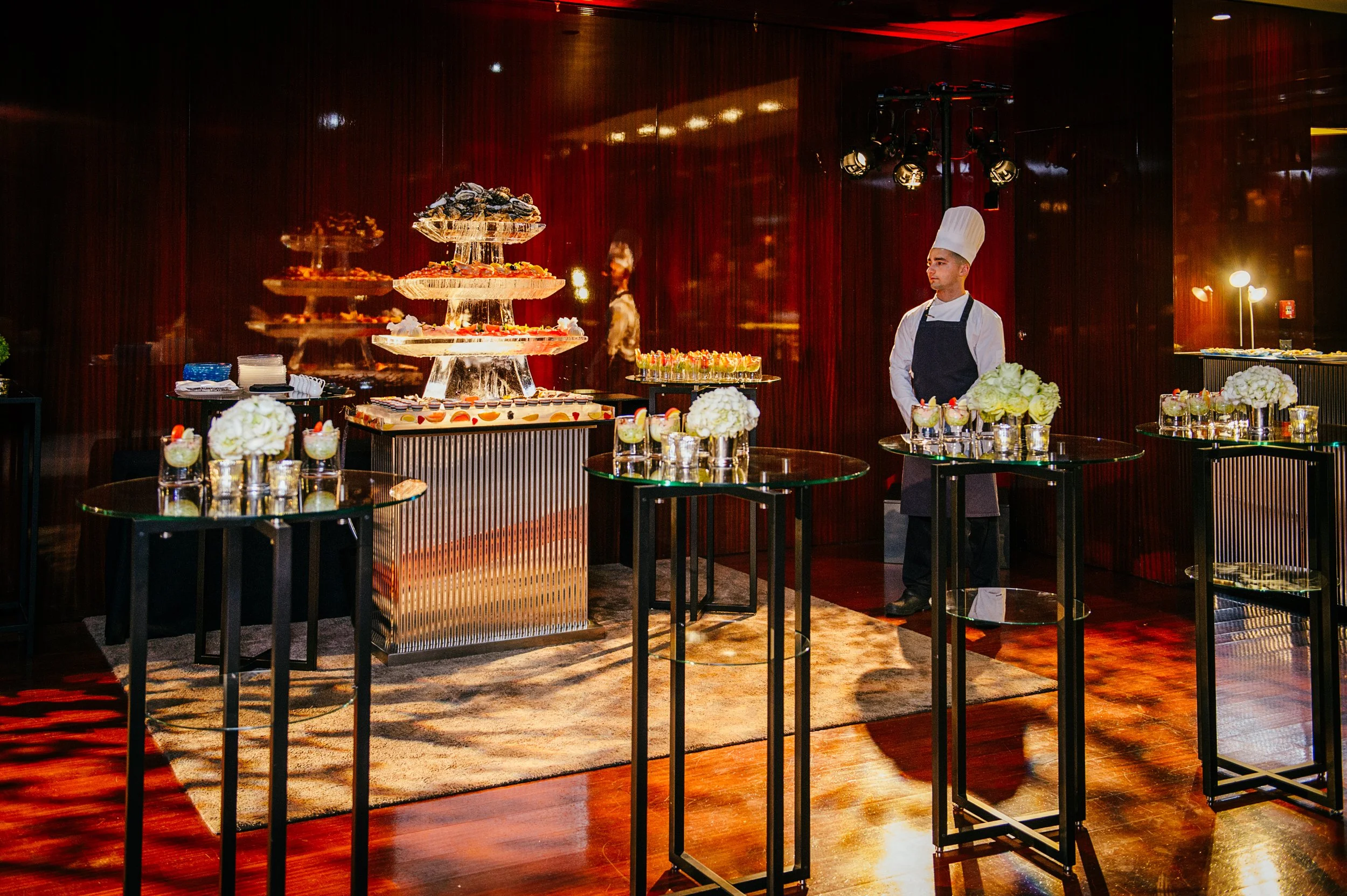 A chef in a tall white hat and black apron standing next to a dessert display at a banquet or event with tiered trays of desserts, glasses with drinks, and floral arrangements, all set against a dark red wooden wall.