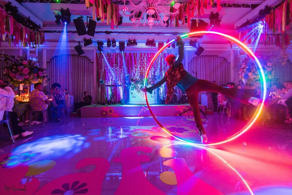 A performer spinning a glowing LED hula hoop in a colorful, illuminated event space with vibrant lighting and floral decorations at Claridge's