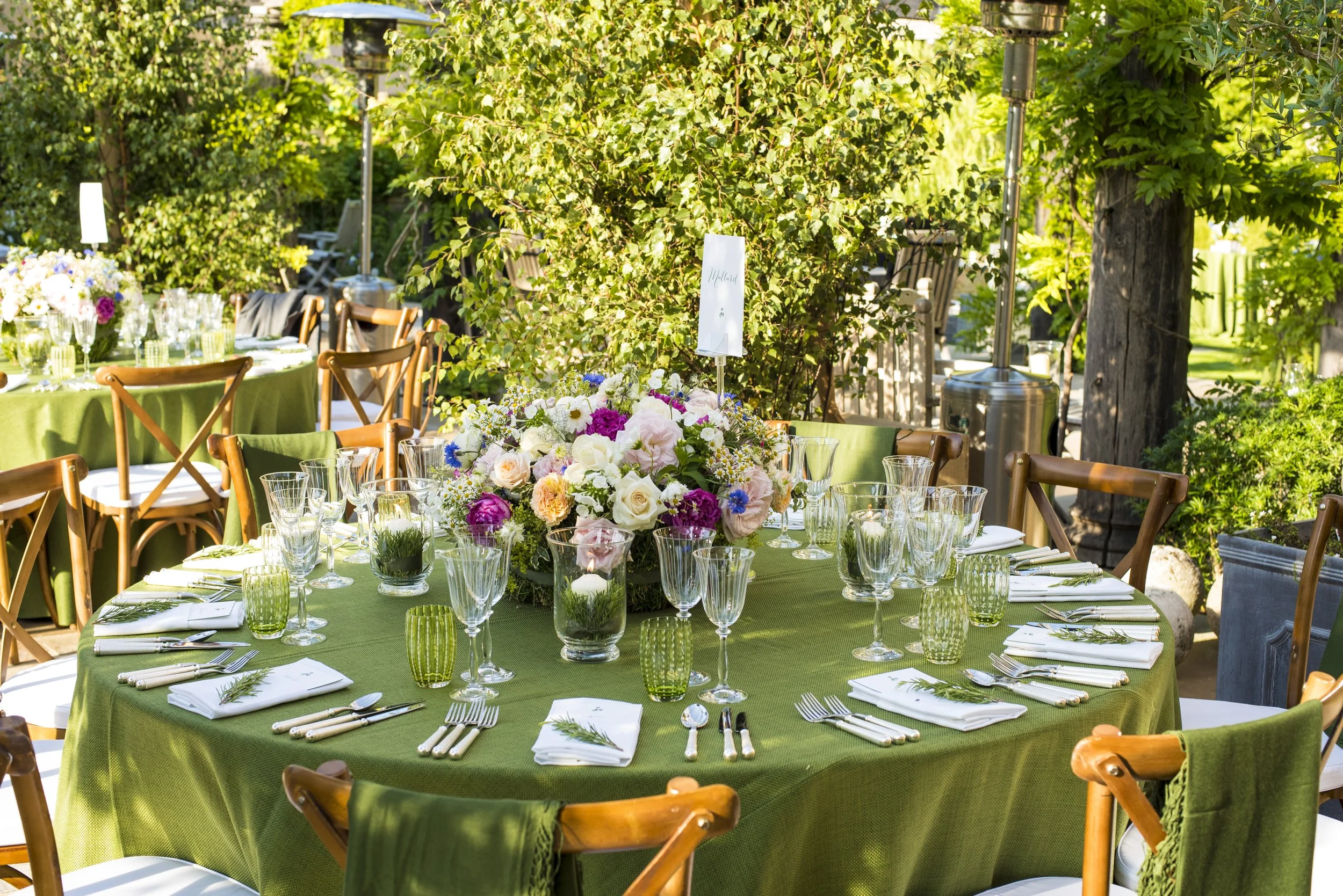 Round table set for an outdoor event with a green tablecloth, floral centerpiece, glassware, and cutlery, surrounded by wooden chairs, with lush green trees and shrubs in the background.