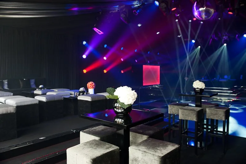 Dimly lit nightclub with seating area and a dance floor illuminated by colorful stage lights.