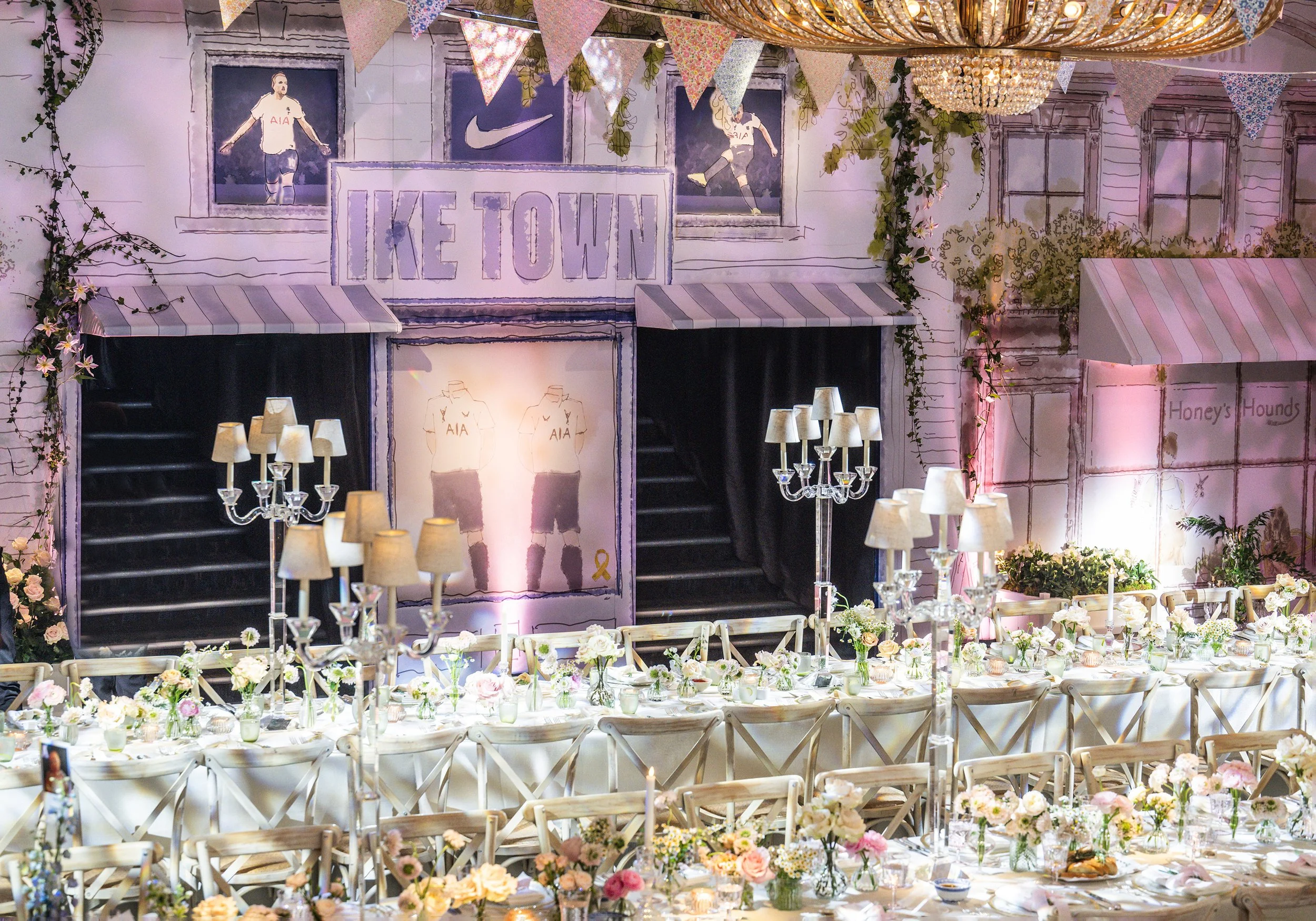 Decorated indoor venue with a long table set for a celebration, featuring numerous small flower arrangements, candle holders, and chairs. The backdrop has an illustration of a house exterior with banners, plants, and portraits of soccer players. Chan