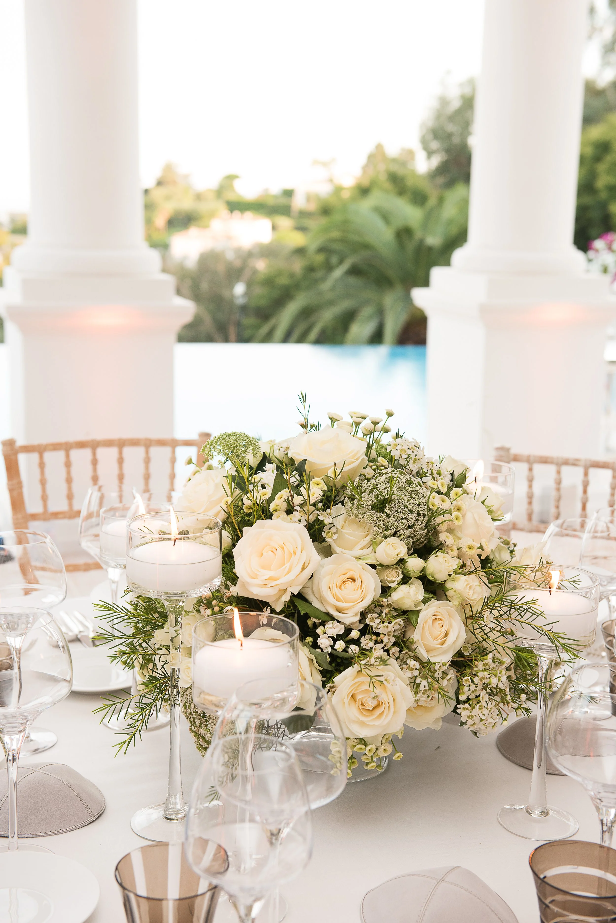 A beautiful table centerpiece with white roses, baby's breath, and greenery, surrounded by lit candles and wine glasses at an elegant event with a view of trees and rooftops in the background.
