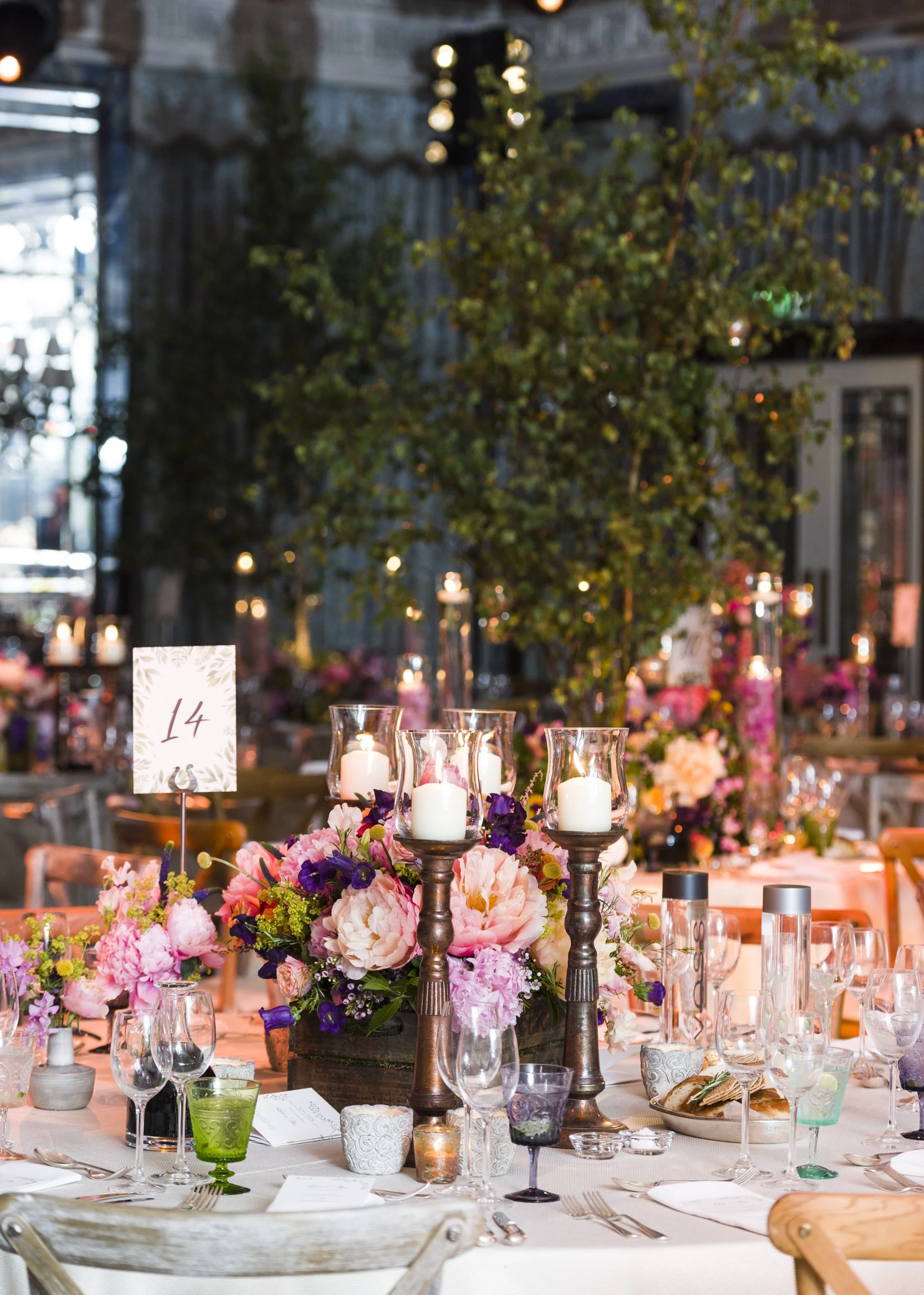 A decorated banquet table with floral centerpieces, candles, and glassware in an indoor setting with dim lighting and greenery hanging from above.