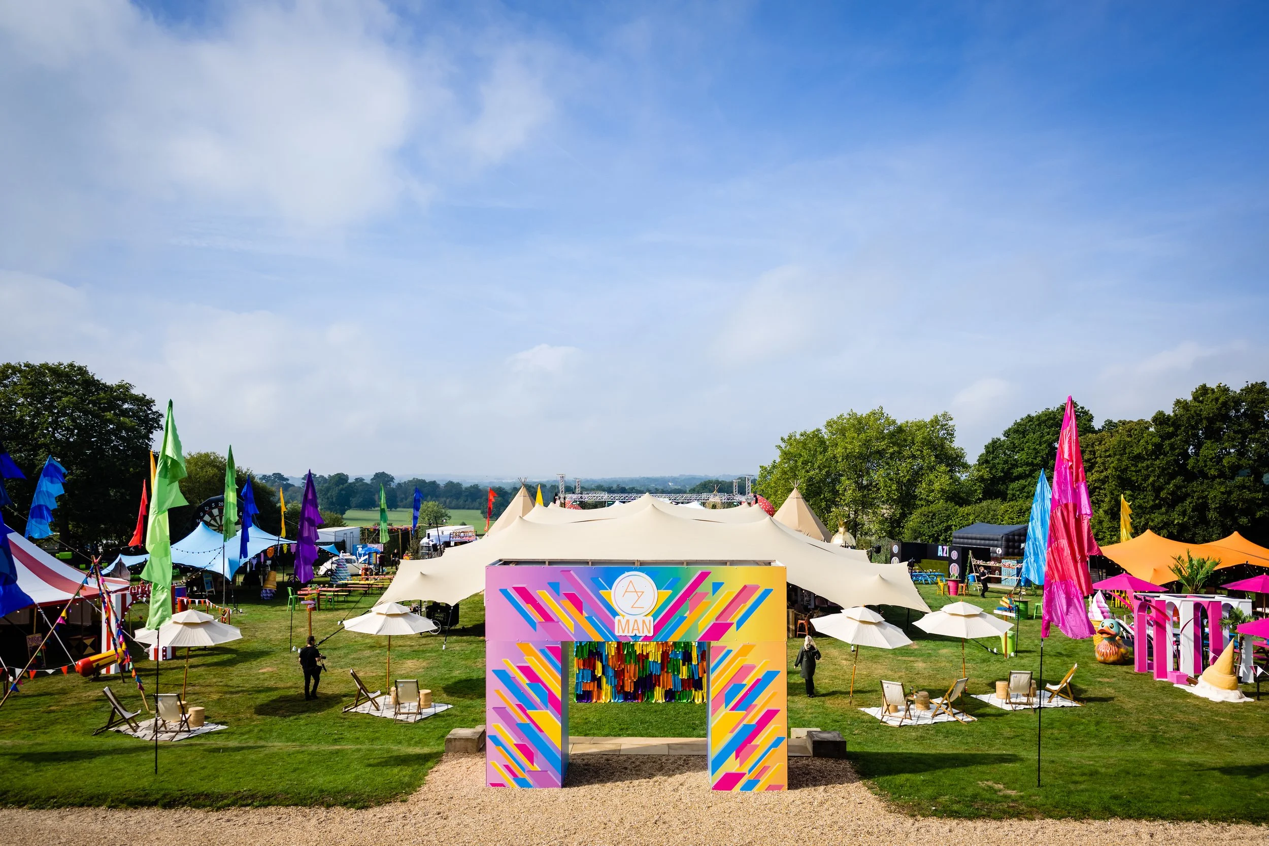 Open field outdoor festival with tents, colorful banners, flags, and a stage under a bright blue sky with some clouds Bar Mitzvah at  Wrotham Park