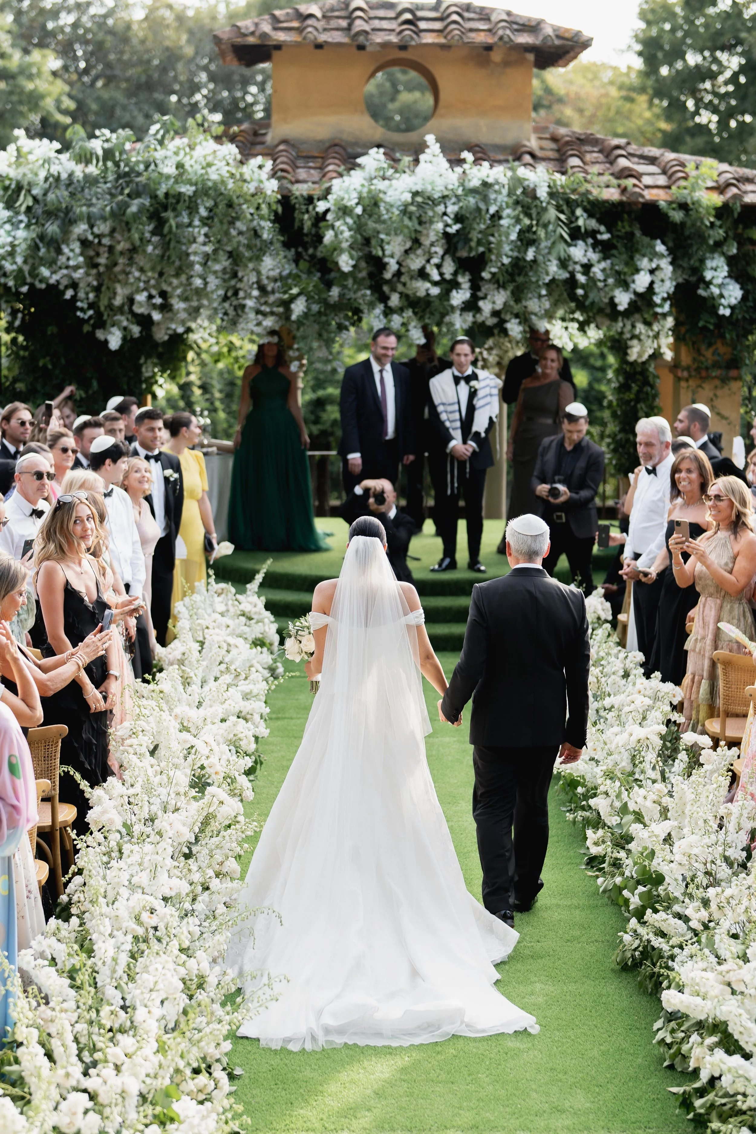 A bride and groom walking down the aisle at an outdoor wedding ceremony with guests on both sides, decorated with white flowers and greenery at Villa Palmieri in Florence