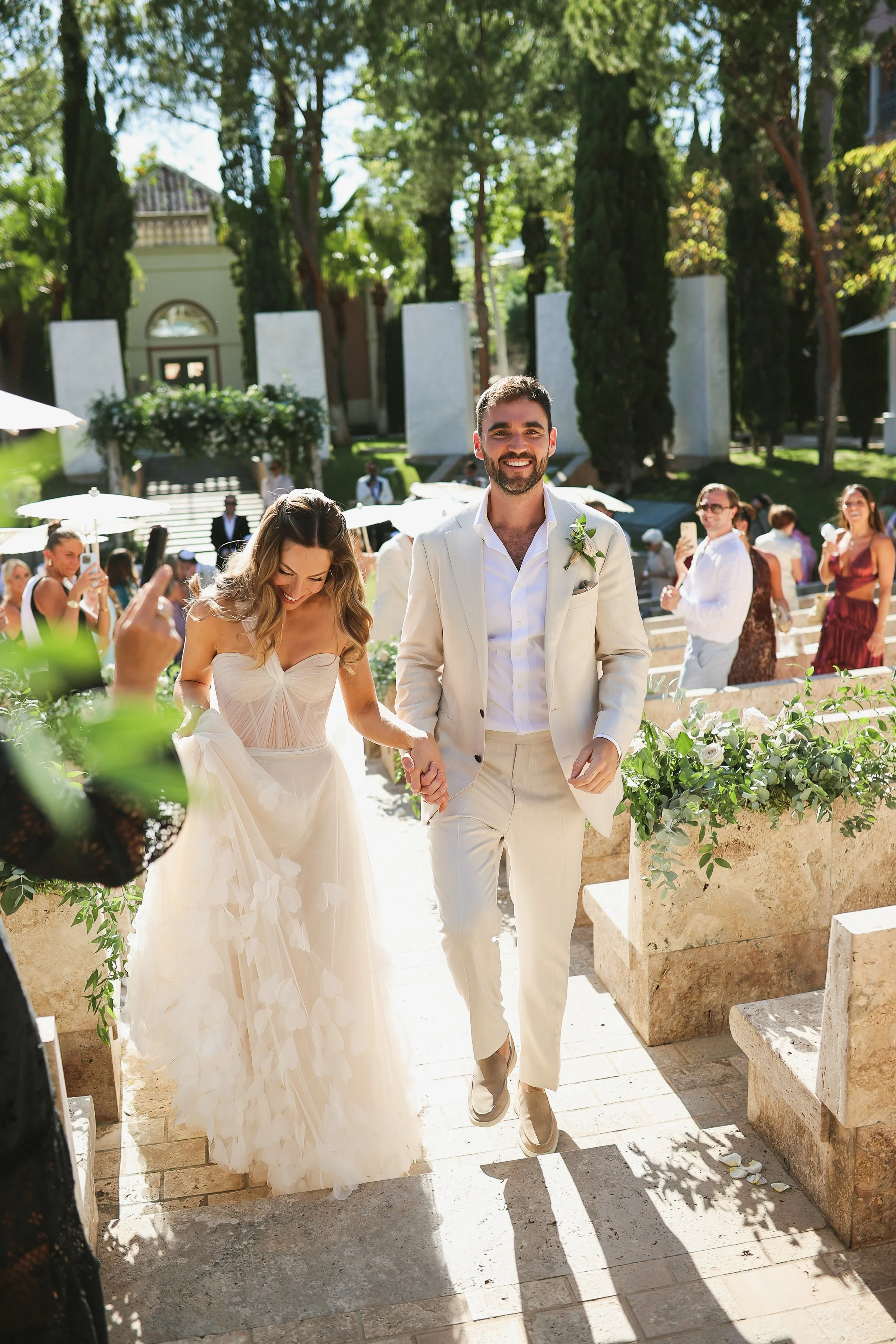 A bride and groom holding hands and walking outdoors during their wedding ceremony, with guests in the background taking photos and smiling, surrounded by greenery and sunny weather.