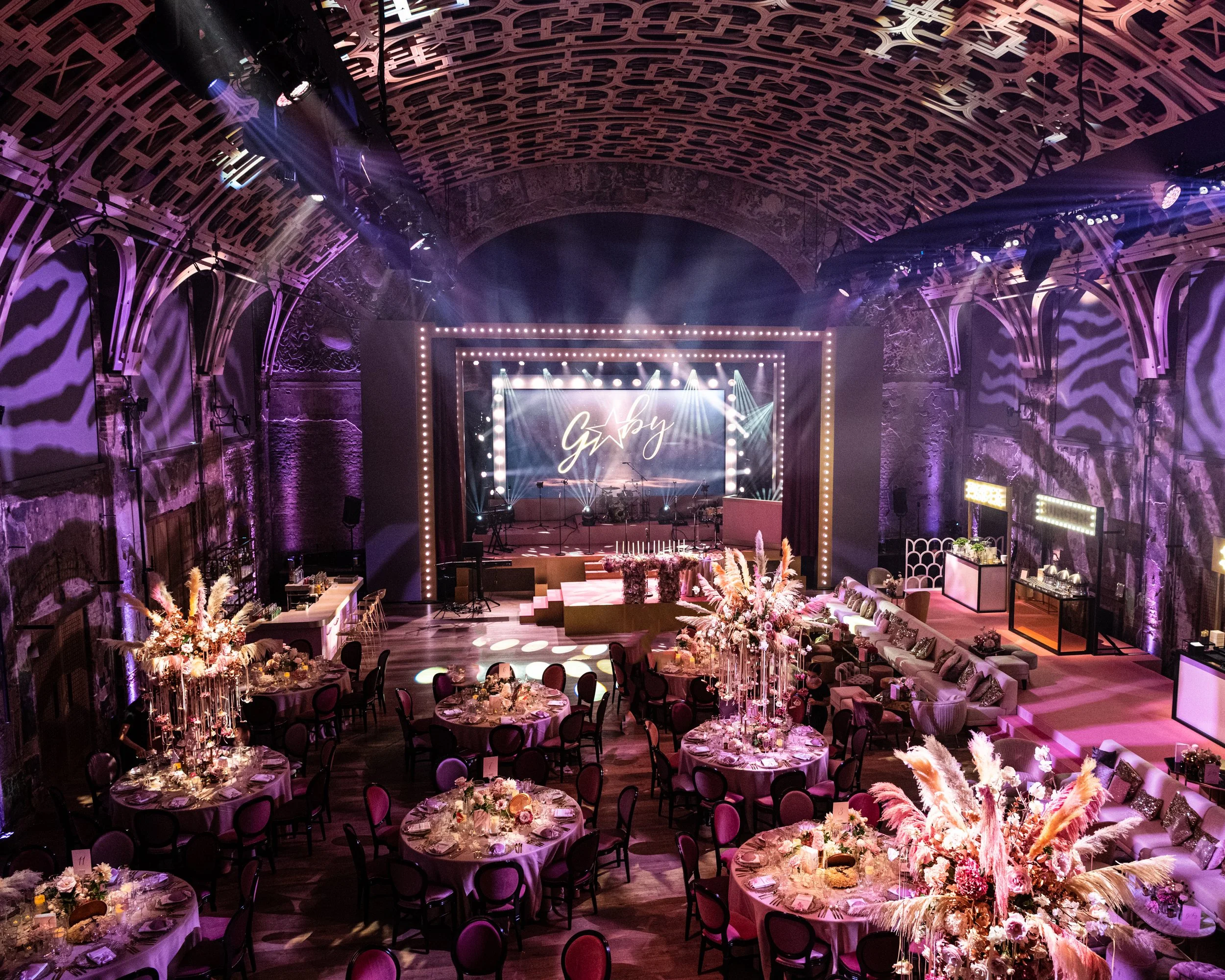 The room features an arched, intricately designed ceiling with purple lighting and a stage with a lit sign that appears to say 'Gaby. BAT MITZVAH AT BATTERSEA ARTS CENTRE