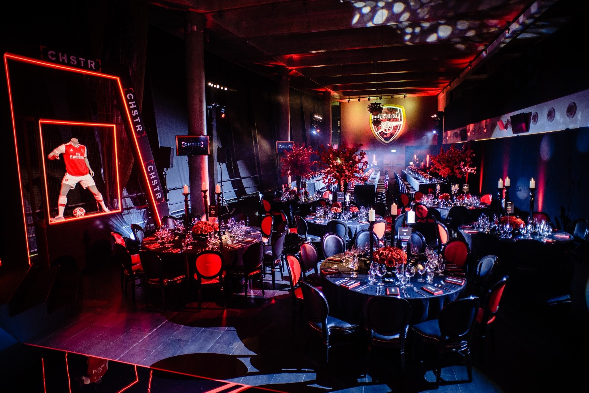 An event space decorated with a football theme, featuring a large Arsenal logo on the wall, a display of a mannequin dressed in an Arsenal football kit, and tables set for a formal dinner with black chairs, red and pink floral centerpieces, and candl