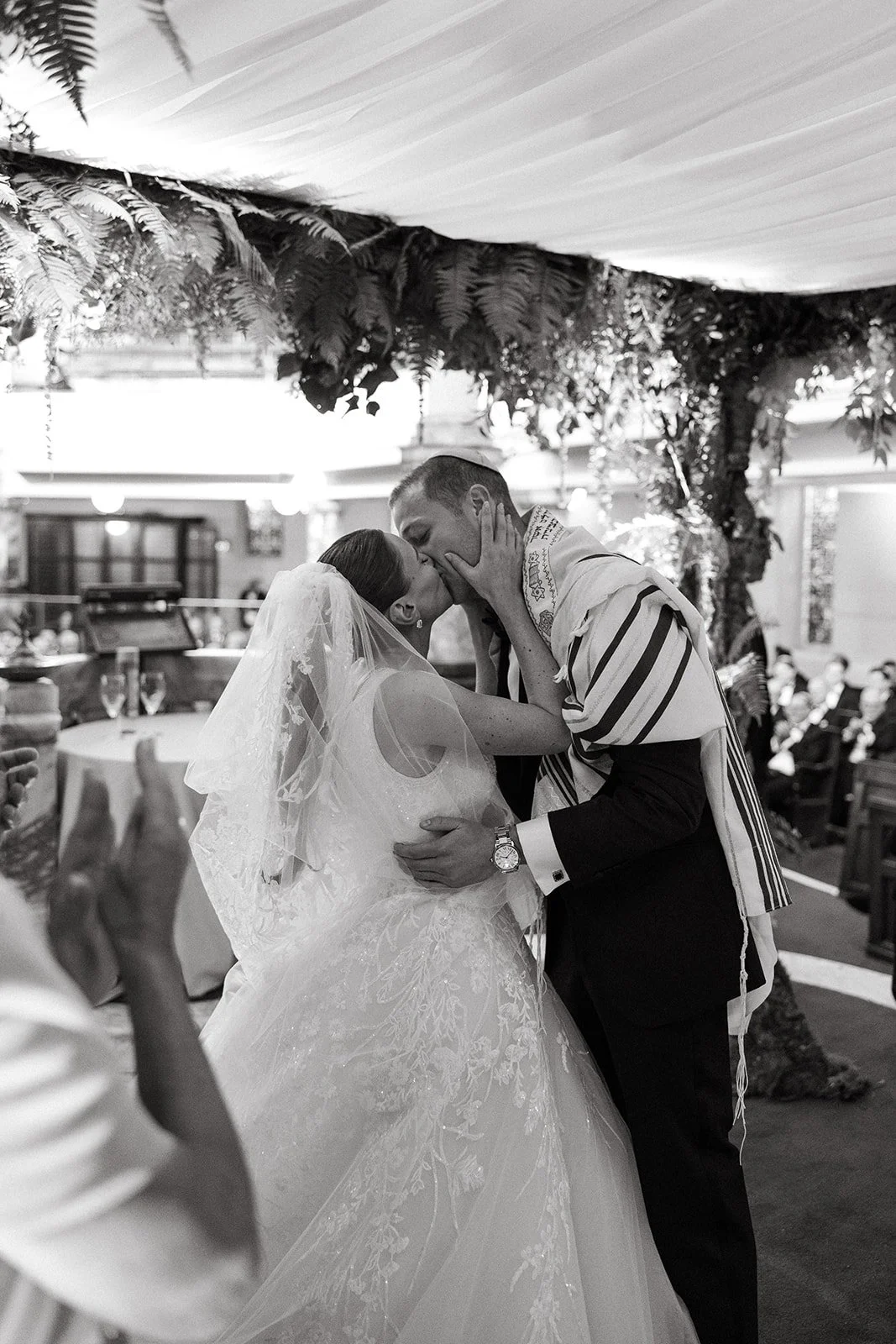 A bride and groom share a kiss during their wedding ceremony, with the bride in a lace wedding dress and veil, and the groom in a kippah and tallit, inside a decorated venue. Jewish wedding