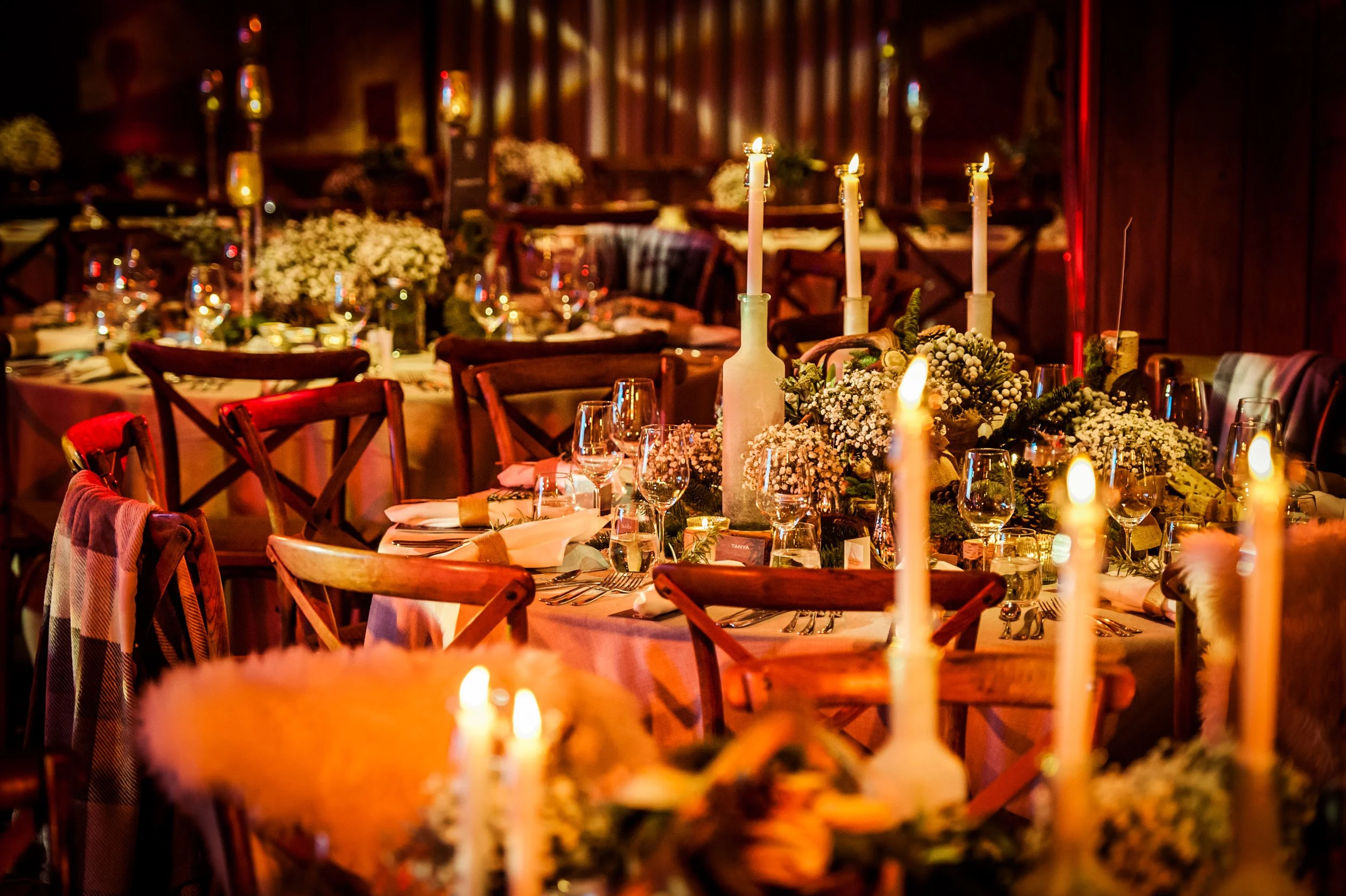 A warmly lit, elegant banquet table decorated with candles, floral centerpieces, and neatly arranged glassware and silverware in a rustic wooden setting.