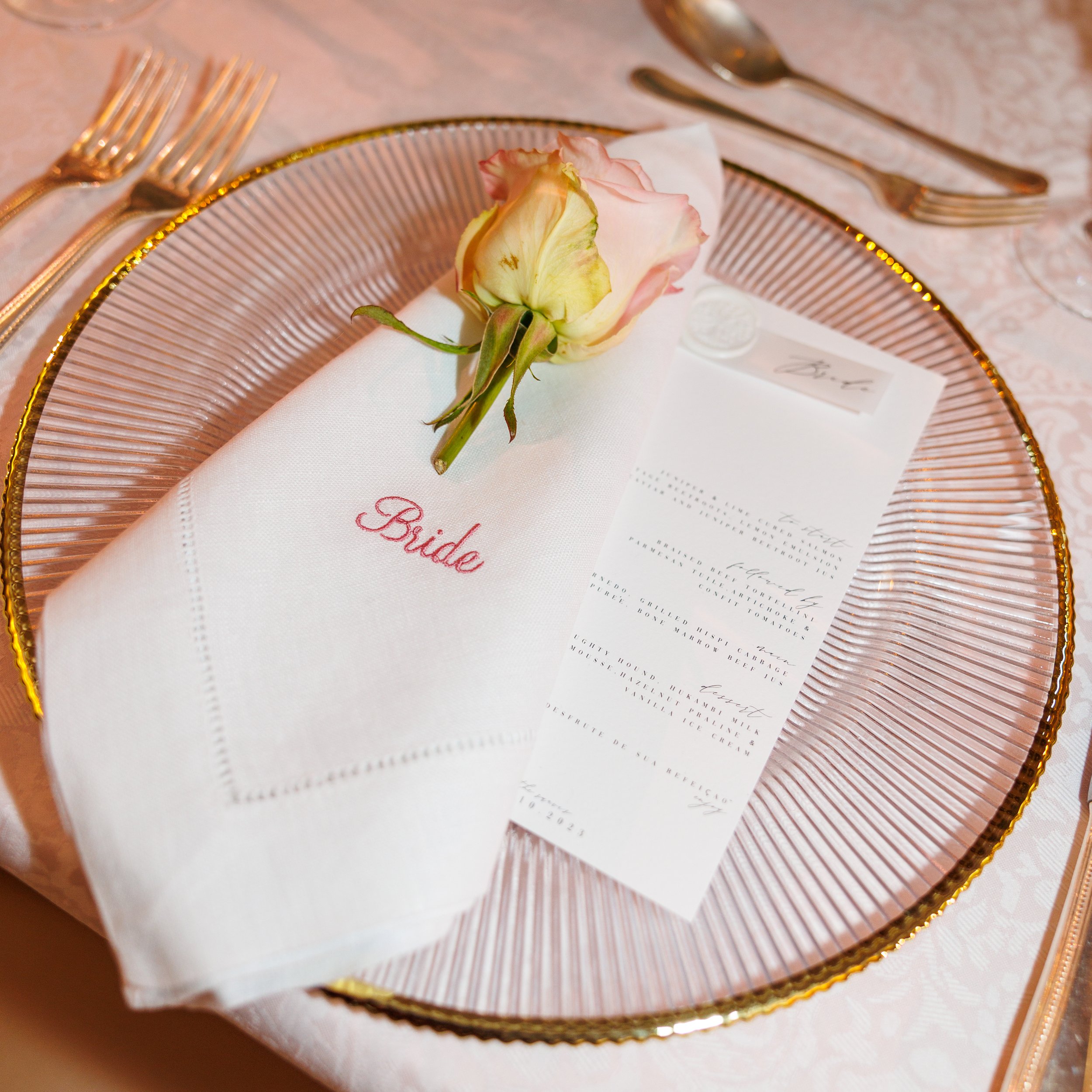 A beige and pink rose placed on a white napkin with the word 'Bride' embroidered in pink, on a decorative pink and gold-rimmed plate. There are gold-colored forks and a spoon set on each side of the plate, and a menu card is partially visible beside 