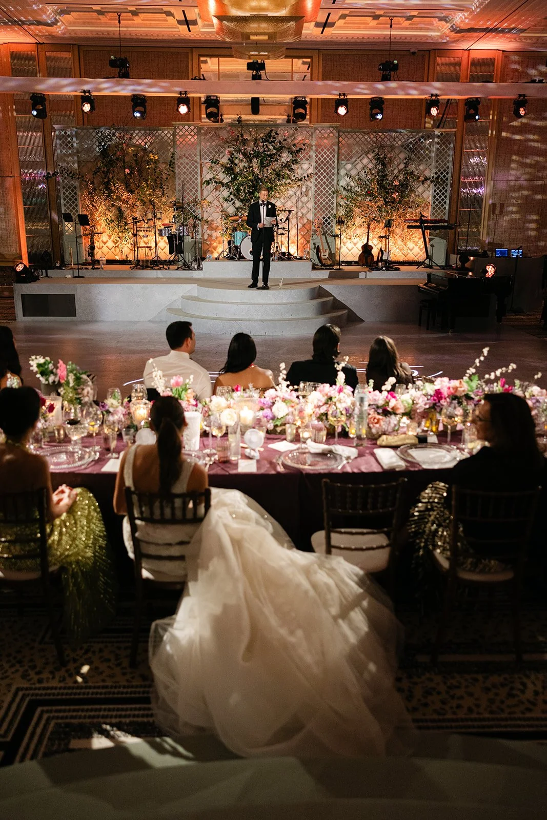 A wedding reception with a bride in a white gown seated at a decorated table, watching a stage with a man in a tuxedo reading from a paper, and musical instruments in the background at The Peninsula London