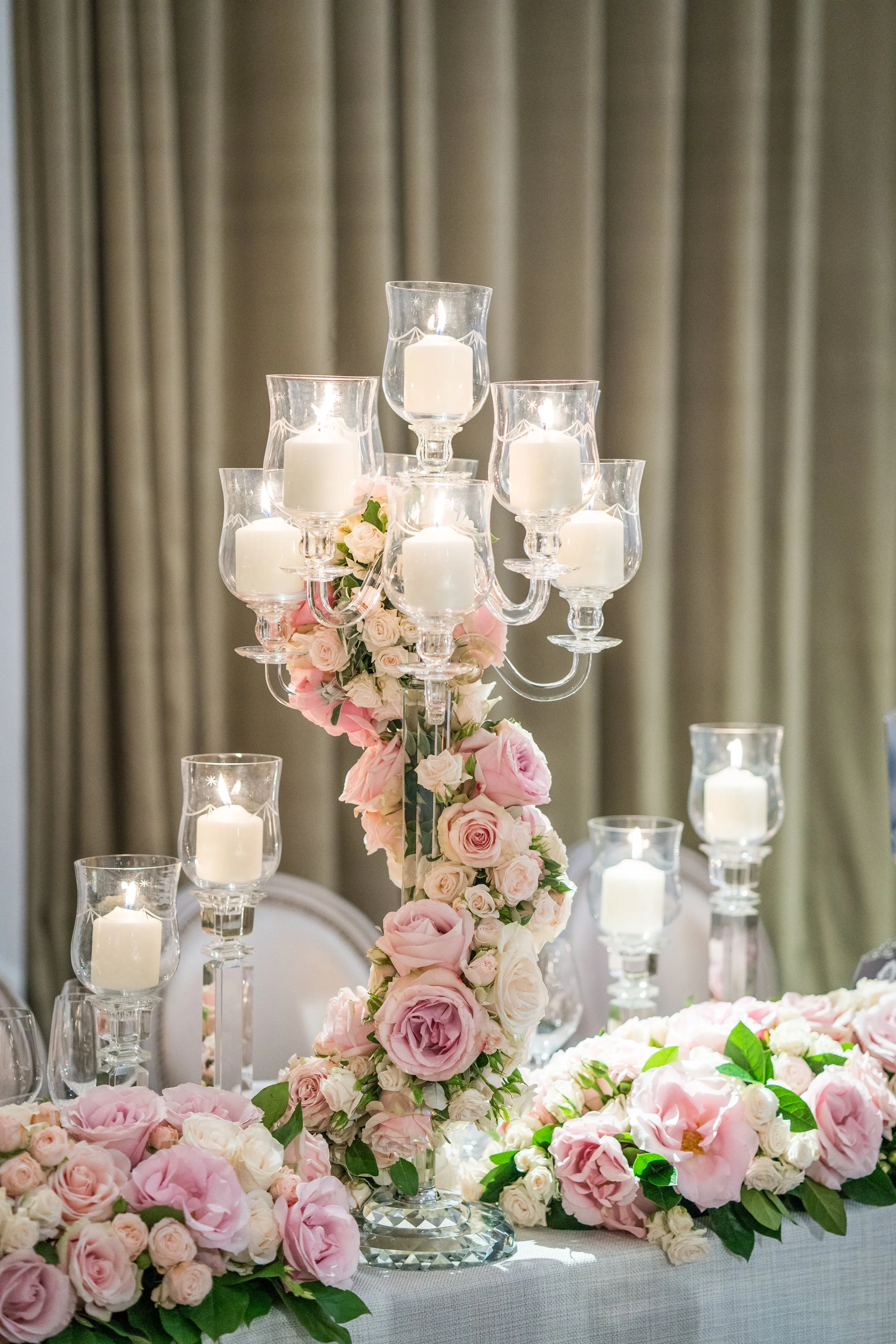 A floral centerpiece with pink and white roses, topped with several lit white candles in glass holders, on a decorated table at an event or celebration.