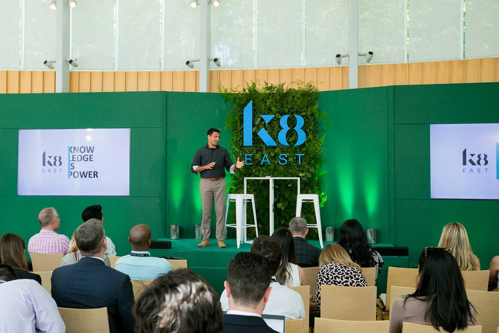 A man presenting on stage at a conference with the logo 'K8 EAST' displayed behind him and a green backdrop with plants. The audience is seated and watching the presentation.