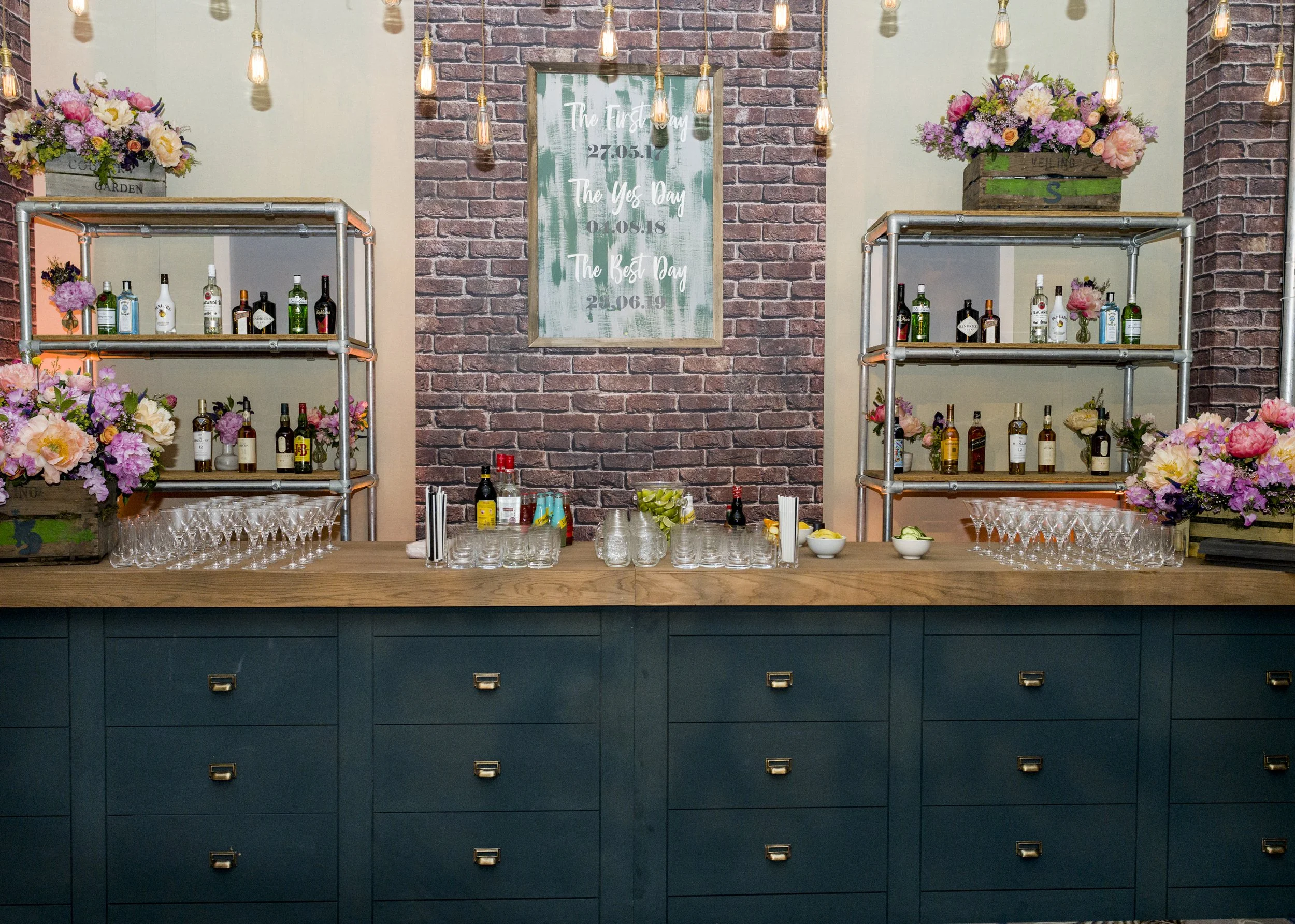 A decorated bar area with bottles of liquor on metal shelves, pink and white flowers, and a wooden sign in the background with wedding dates and phrases.
