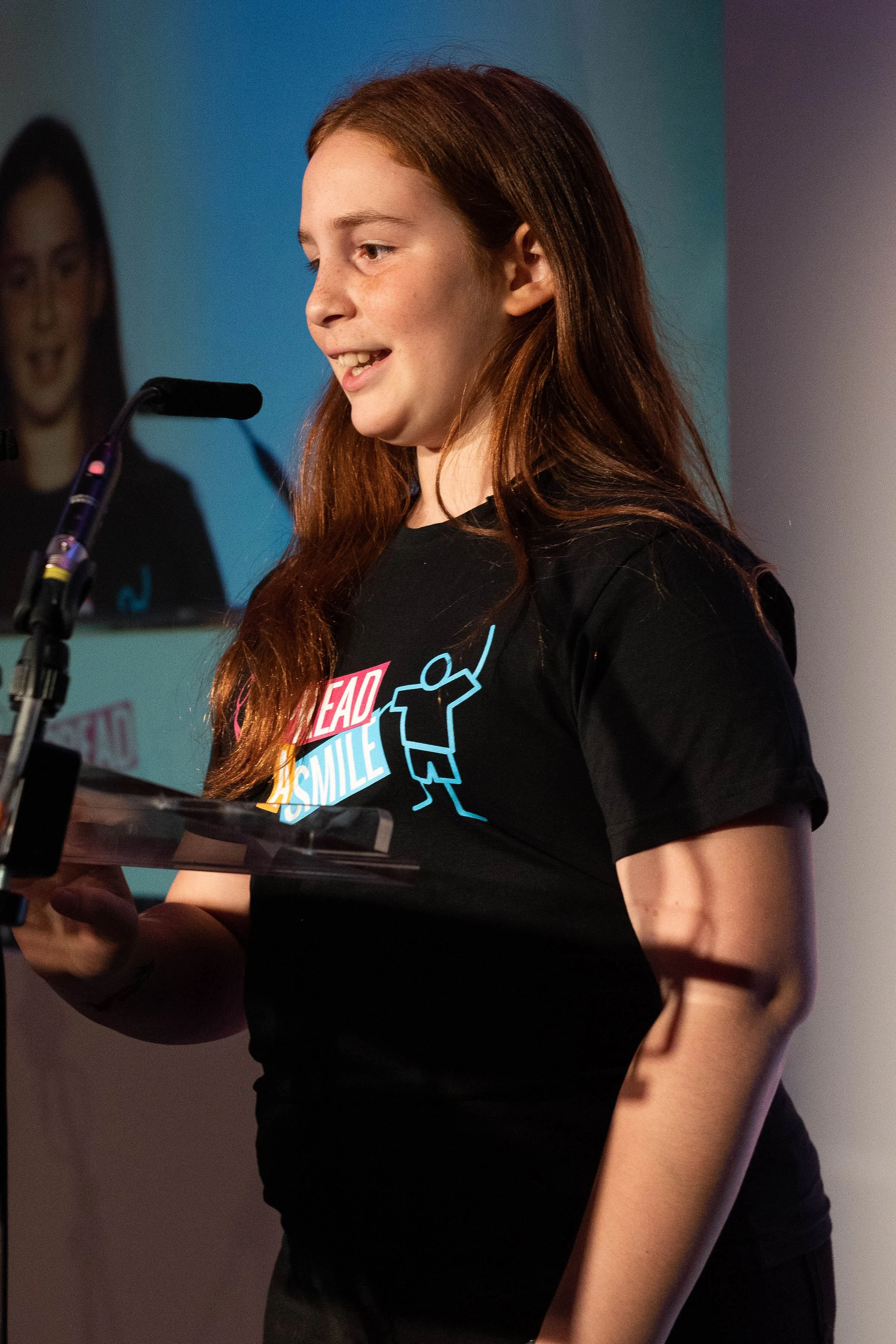 Young woman with long red hair speaking into a microphone at a podium, wearing a black t-shirt with colorful text and graphics.