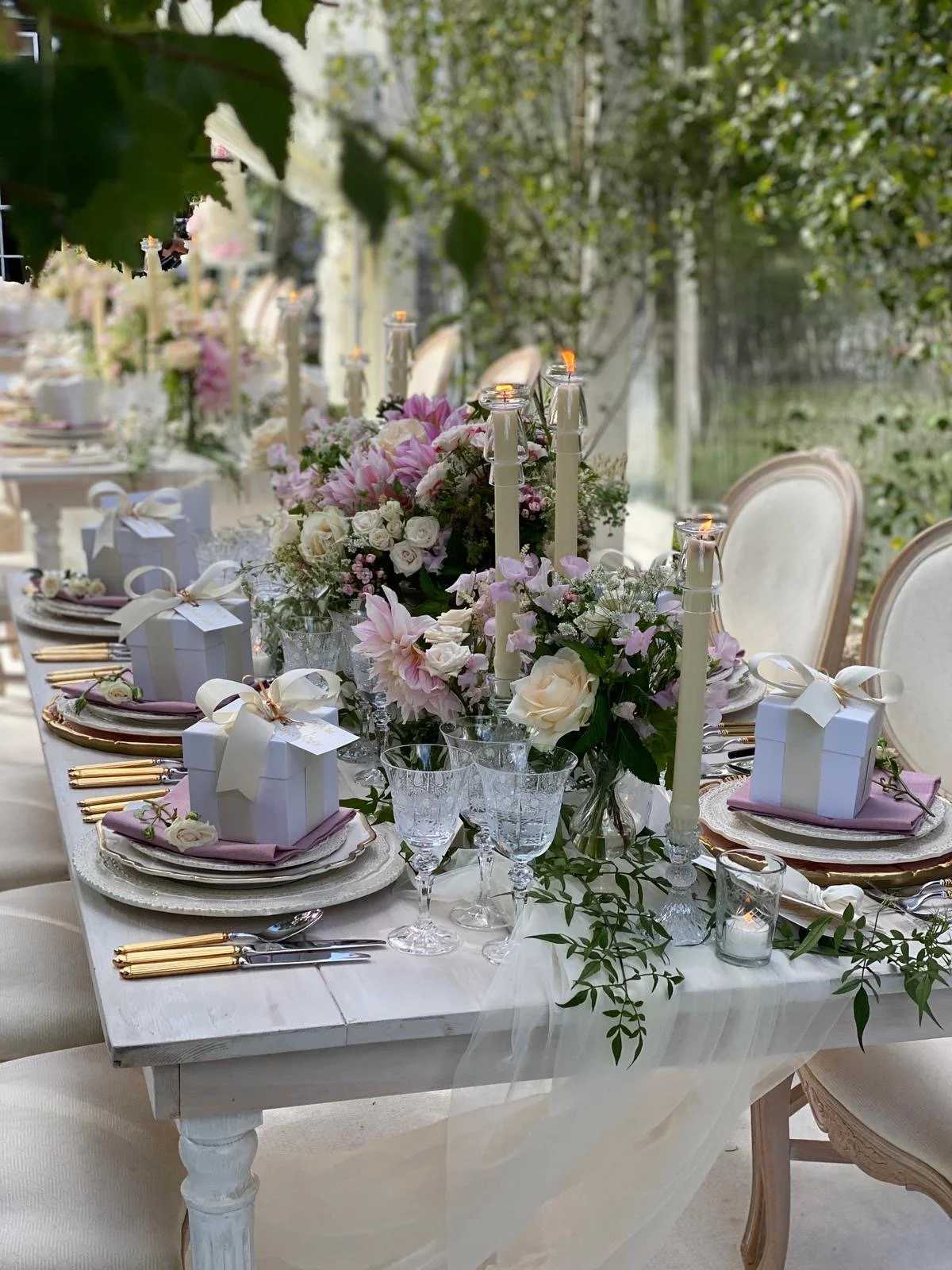Elegant outdoor wedding table decorated with pink and white flowers, candles, glassware, and gift boxes.