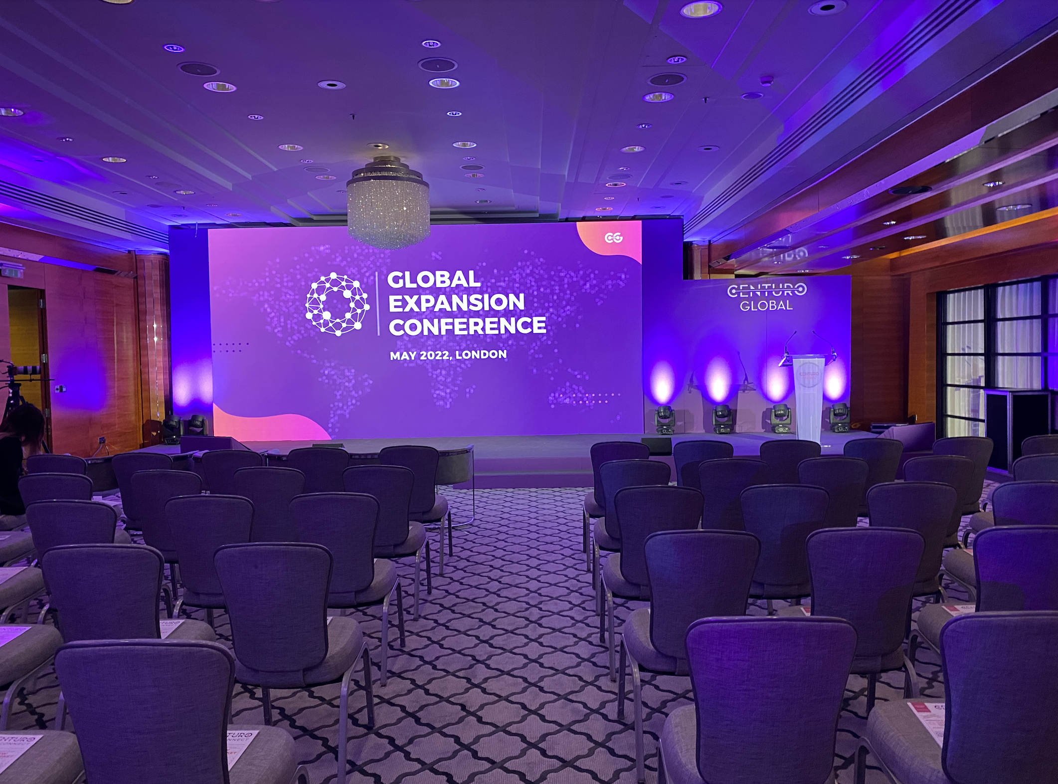 Conference room with rows of chairs facing a large purple screen displaying the words "Global Expansion Conference" and "May 2022, London". The room features a chandelier, purple lighting, and a wooden interior.