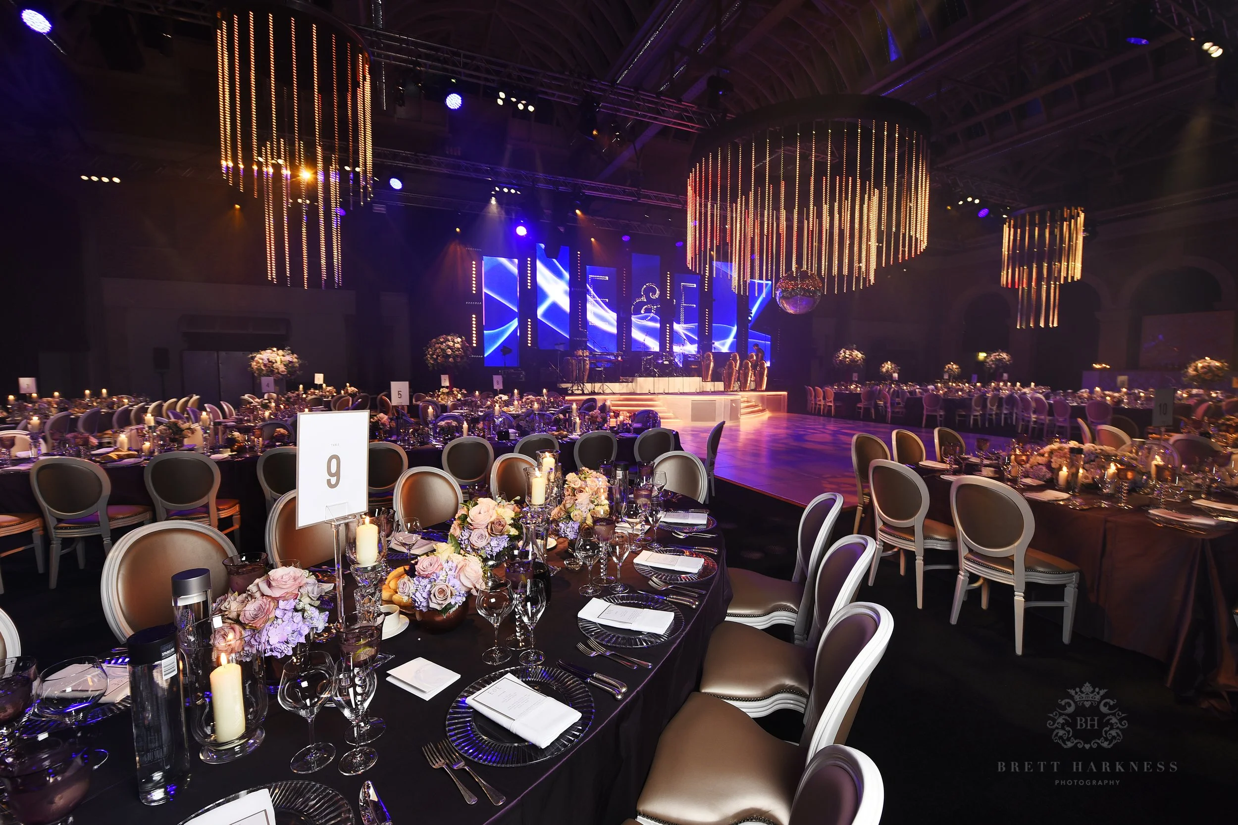 A banquet hall decorated for a formal event with round tables, floral centerpieces, and candles, a stage with colorful lighting and large digital screens, and several chandeliers hanging from the ceiling.