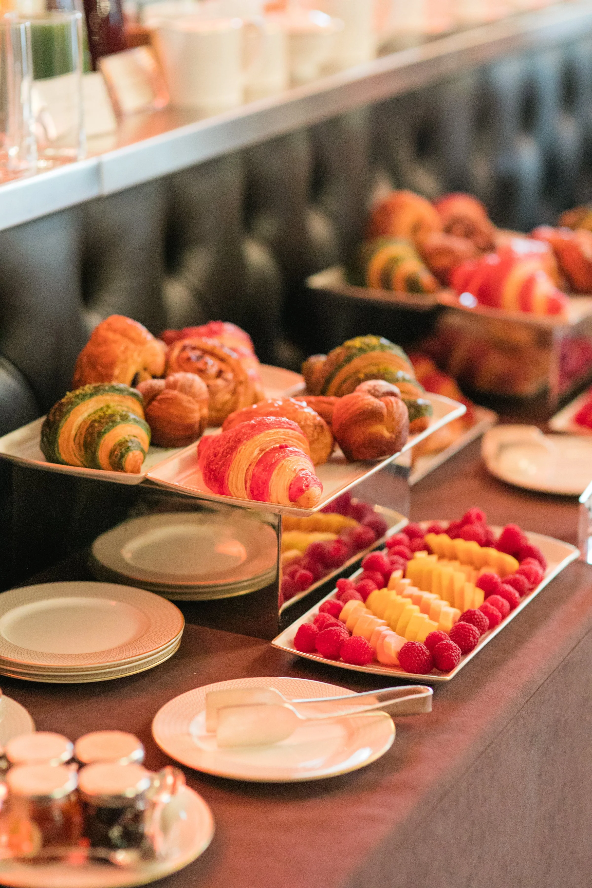 A buffet table with assorted pastries including croissants wrapped in various colored laminations, and a tray of fresh fruit, such as raspberries and sliced melons, served on white rectangular plates.