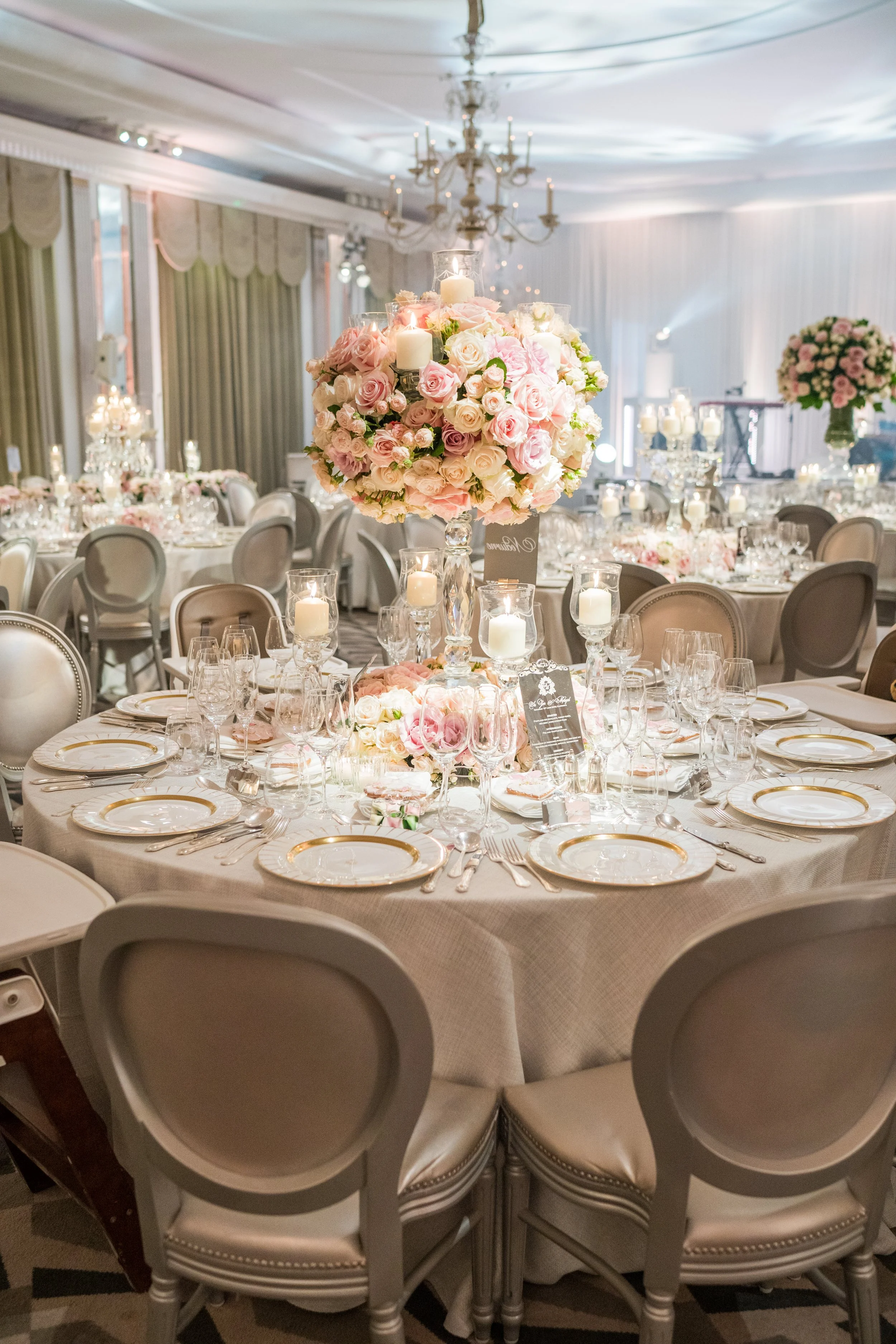 Elegant banquet hall decorated with large floral centerpieces of pink and white roses, surrounded by lit candles on a round table set with white tablecloths, fine china, glassware, and silverware, with additional tables and floral arrangements in the