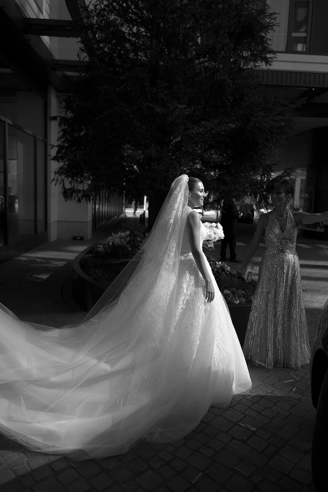 A bride in a wedding gown with a long veil smiling as she interacts with a woman in a beaded dress, outside near a large tree at The Peninsula London