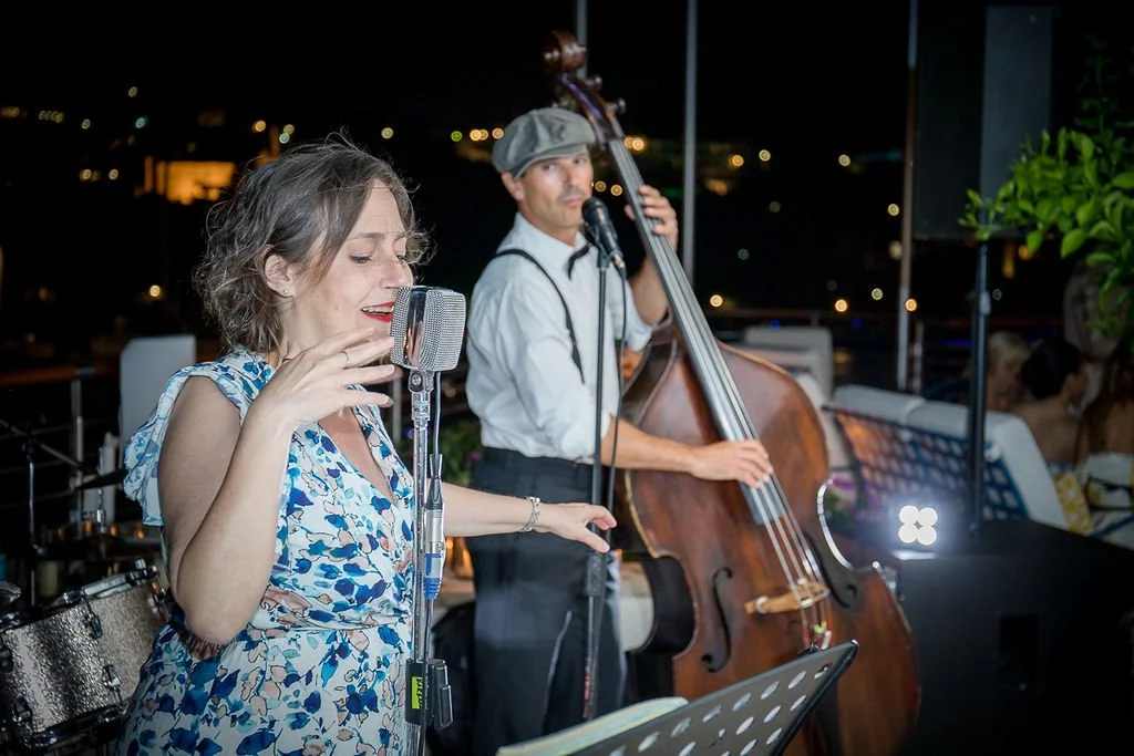 A woman singing into a microphone with a jazz band playing behind her at night on an outdoor terrace.