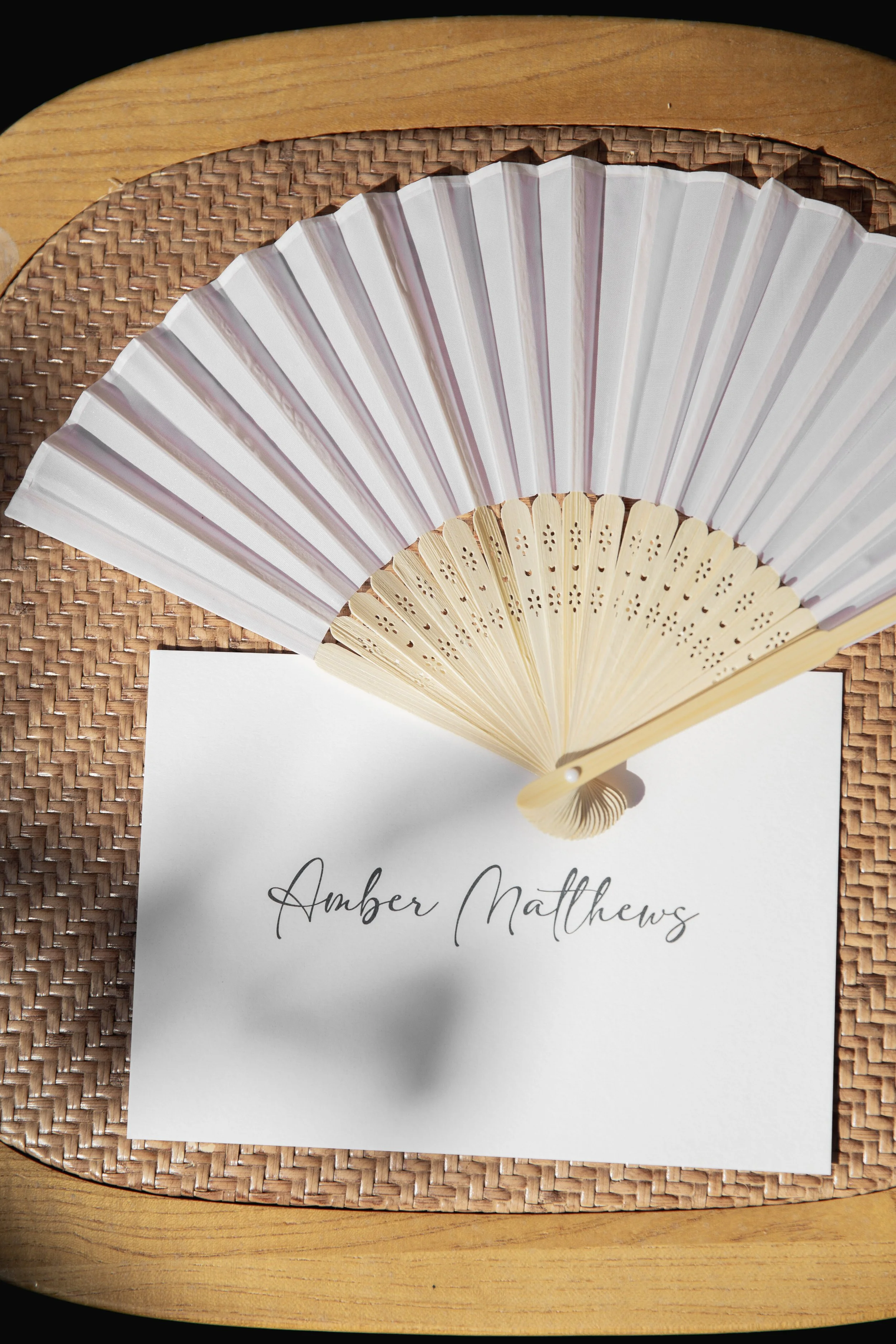 A white paper with the names written in elegant cursive font, placed on a woven placemat with a wooden surface underneath. A decorative foldable hand fan with light-colored bamboo and white fabric is positioned above the paper.