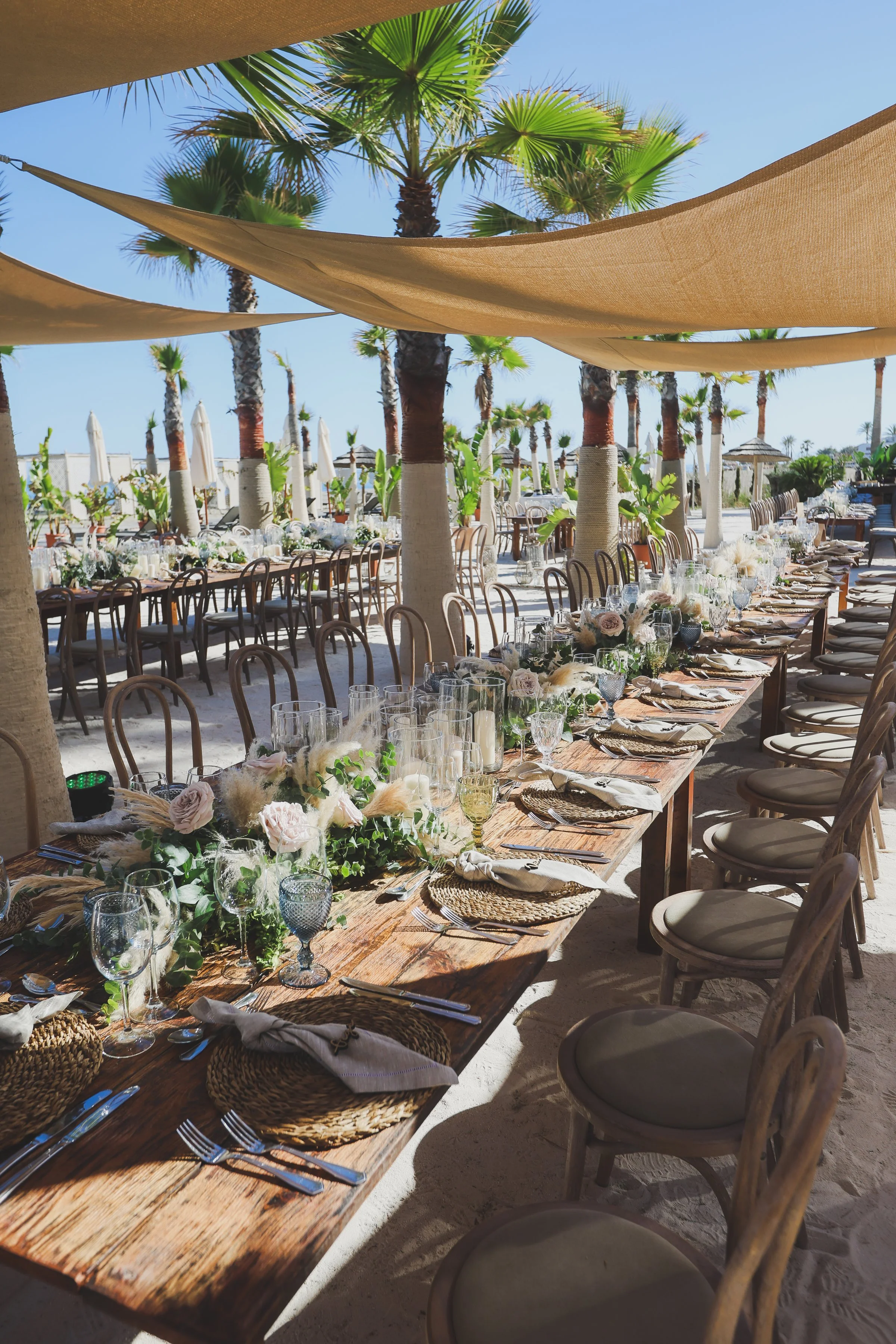 Long outdoor dining table set for a celebration, decorated with flowers, candles, glassware, and linen napkins, under beige sunshades with palm trees in the background on a sunny beach.