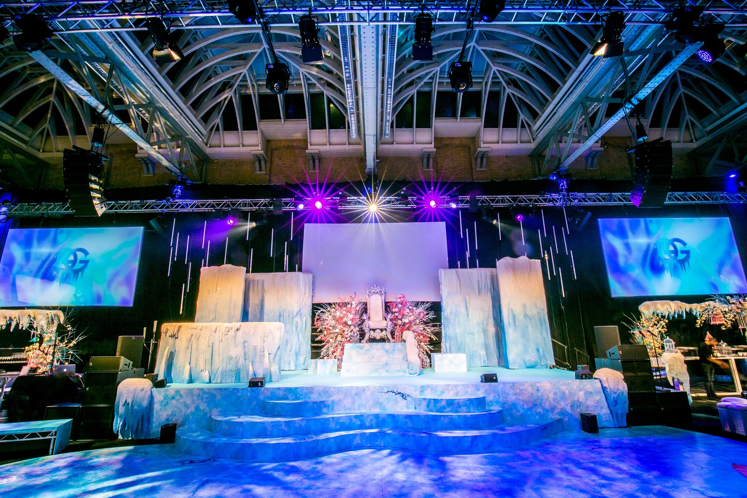 Event venue stage with decorative flowers, draped fabric, and lighting, set up for a celebration or wedding in an indoor space with a high, arched ceiling and stage lighting. ICE PALACE BAT MITZVAH at Old Billingsgate 