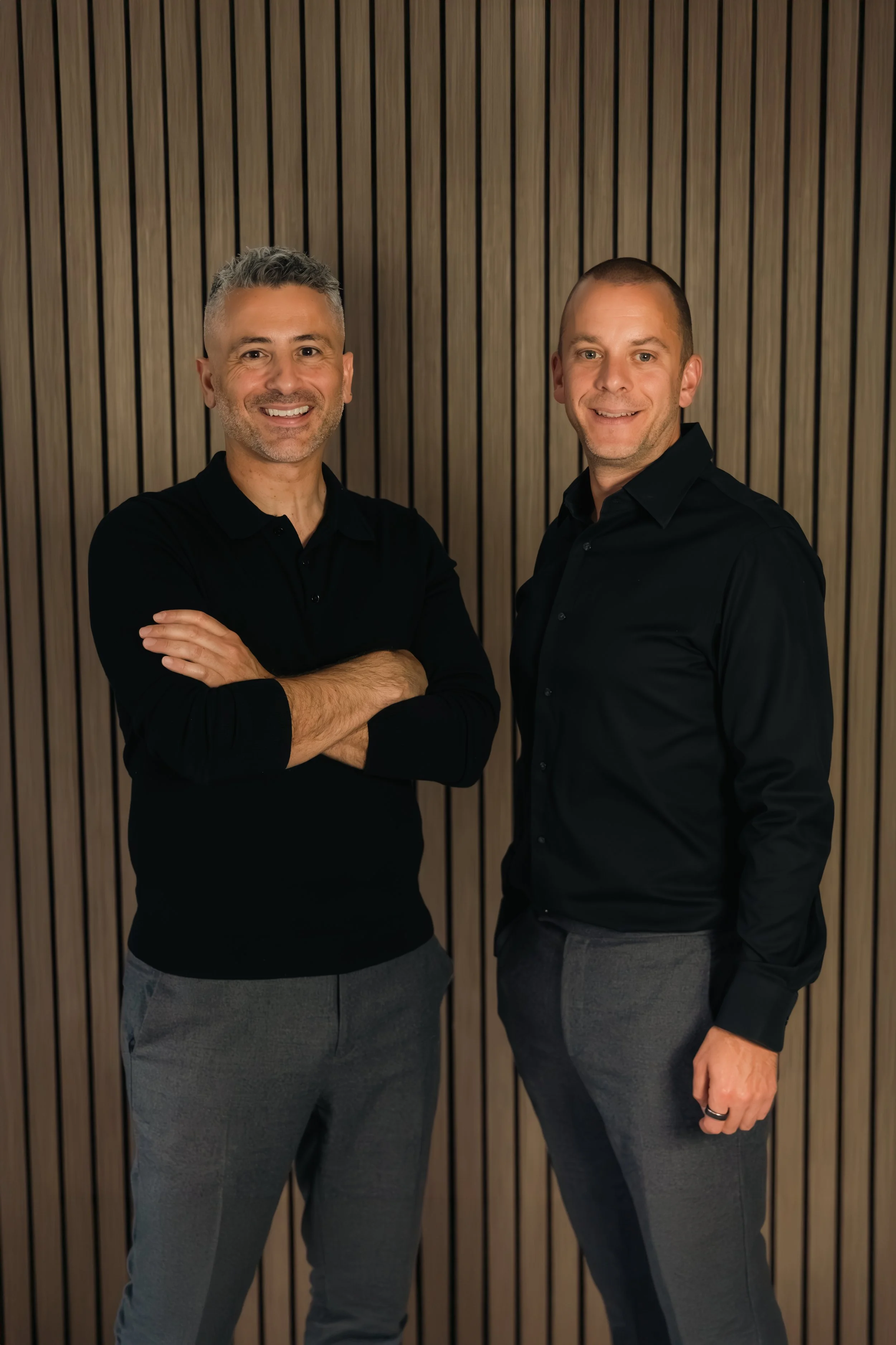 Laurie Weitzkorn and Stas Anastasiou in black shirts and gray pants standing in front of a wooden slat wall, smiling at the camera.