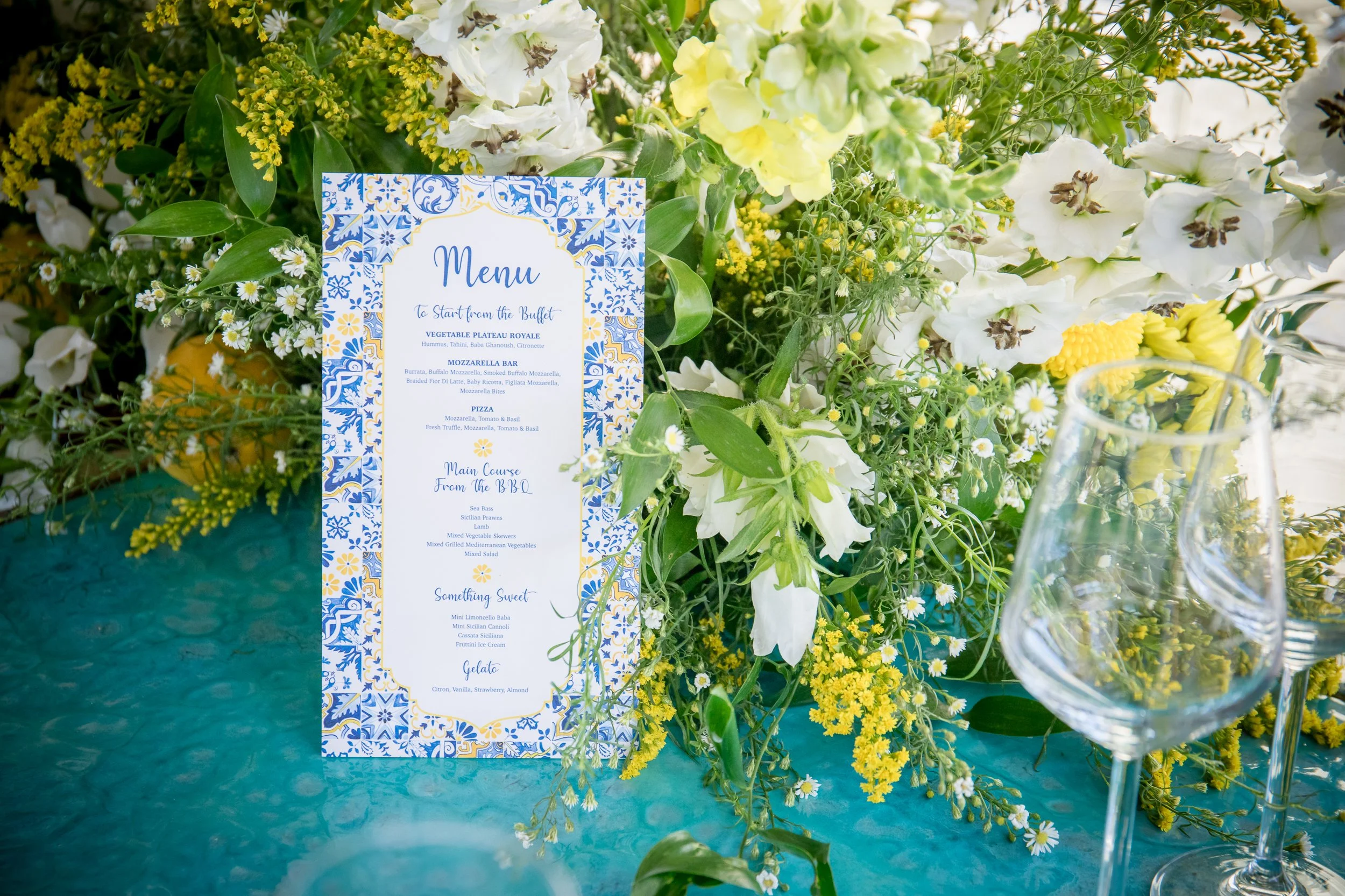 A colorful menu card with a blue and yellow pattern in a floral design, placed among white, yellow, and green flowers on a table.