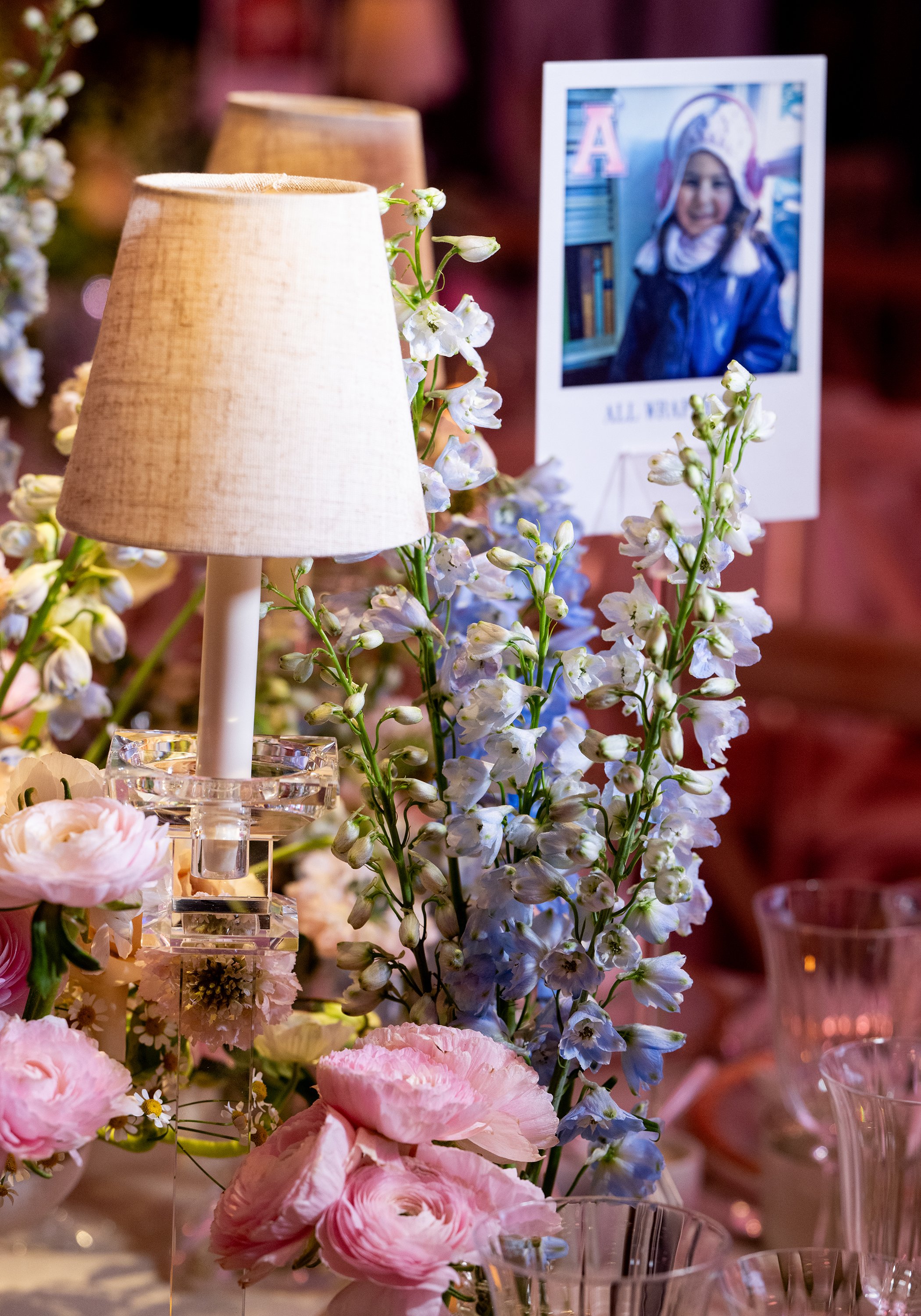 A decorative table arrangement of soft pink and white flowers, including ranunculus, with a small beige lampshade on a glass base and a photo of a smiling girl in a blue jacket and white hat in the background, surrounded by other floral decor.