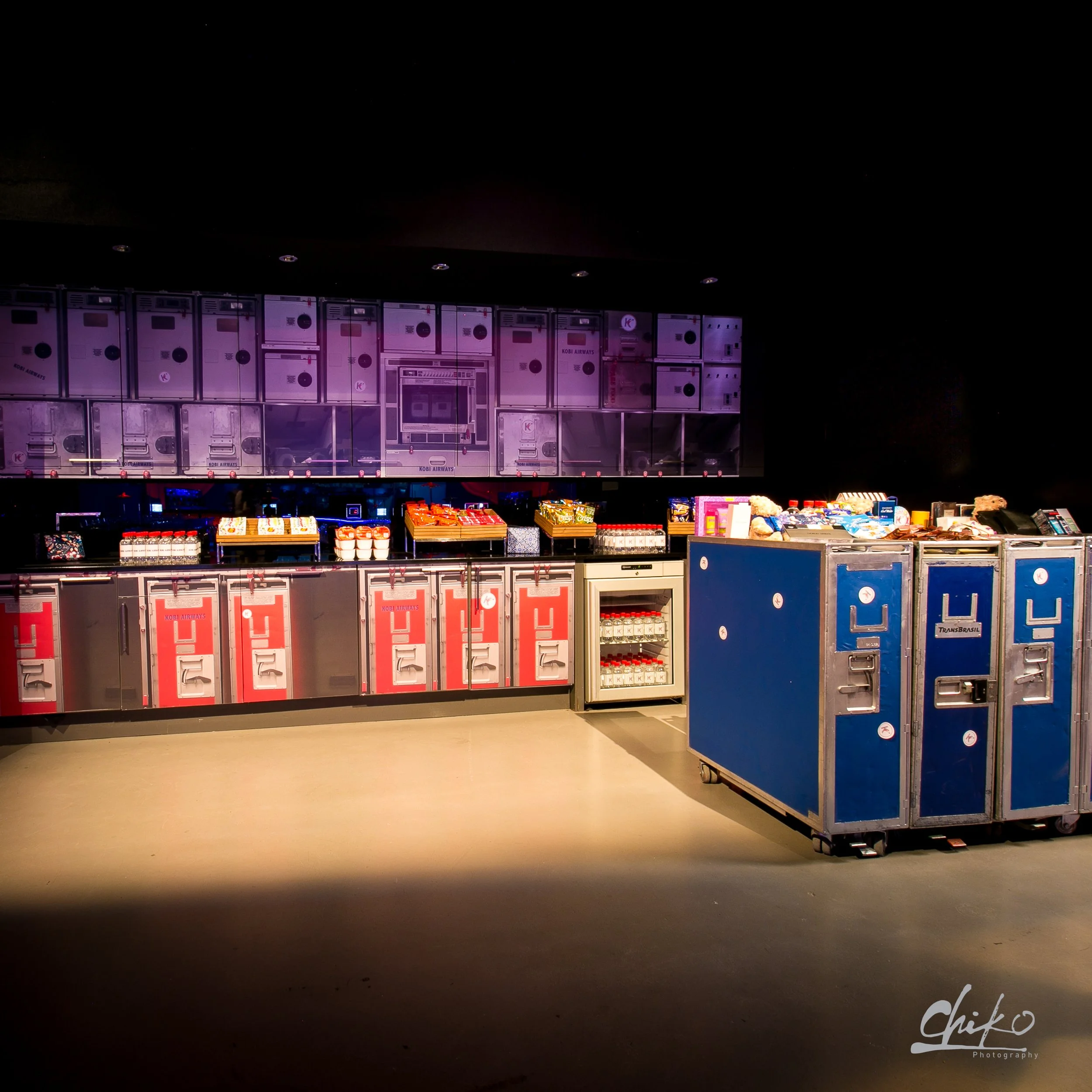 A row of vending machines or lockers with a dark background, illuminated by dim lighting, and various snacks or items on a table in front.