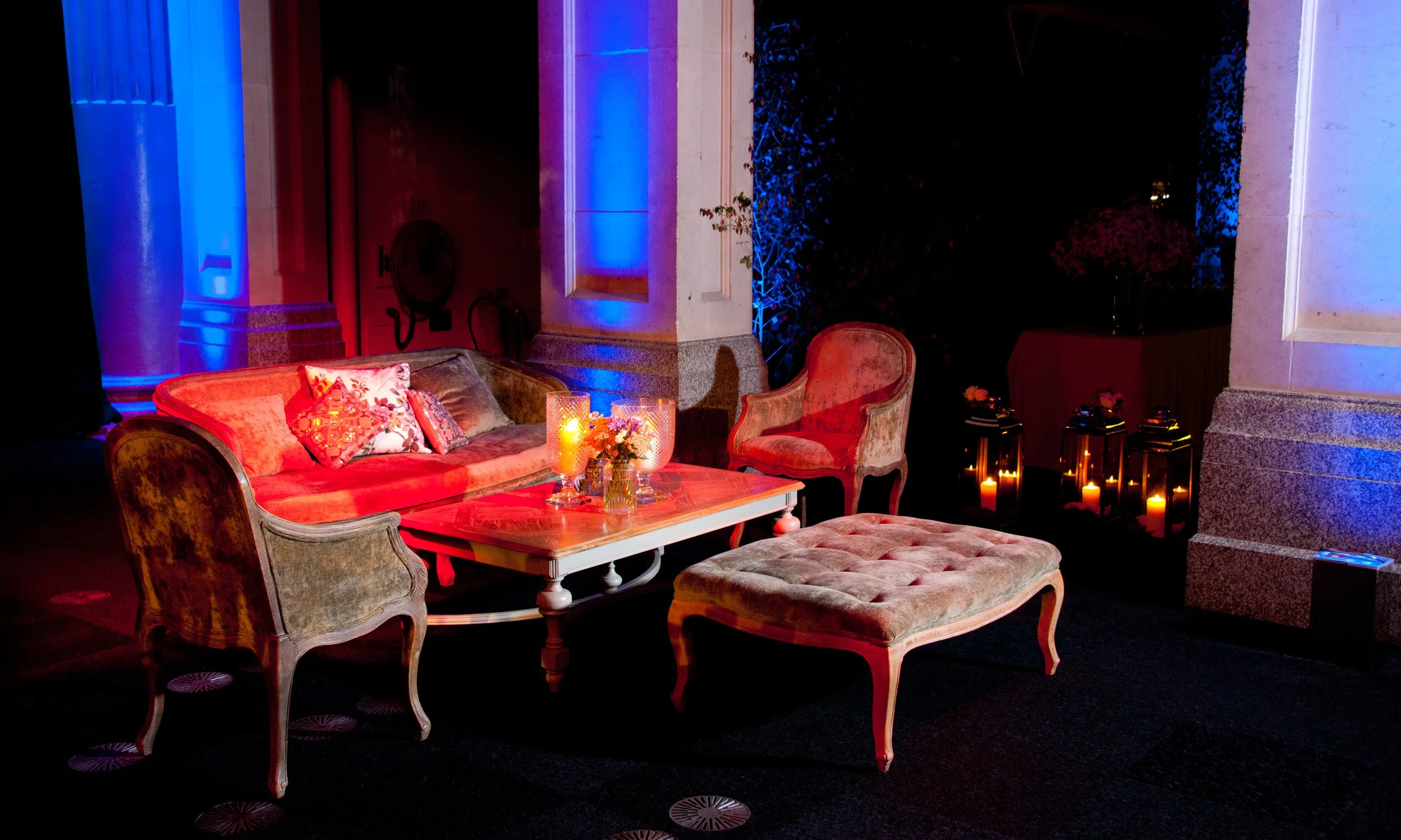 A cozy indoor seating area with vintage-style chairs and a sofa arranged around a coffee table with candles and flowers. The room is dimly lit with colorful blue and red lighting, with some candles in lanterns on the floor and decorative pillows on t