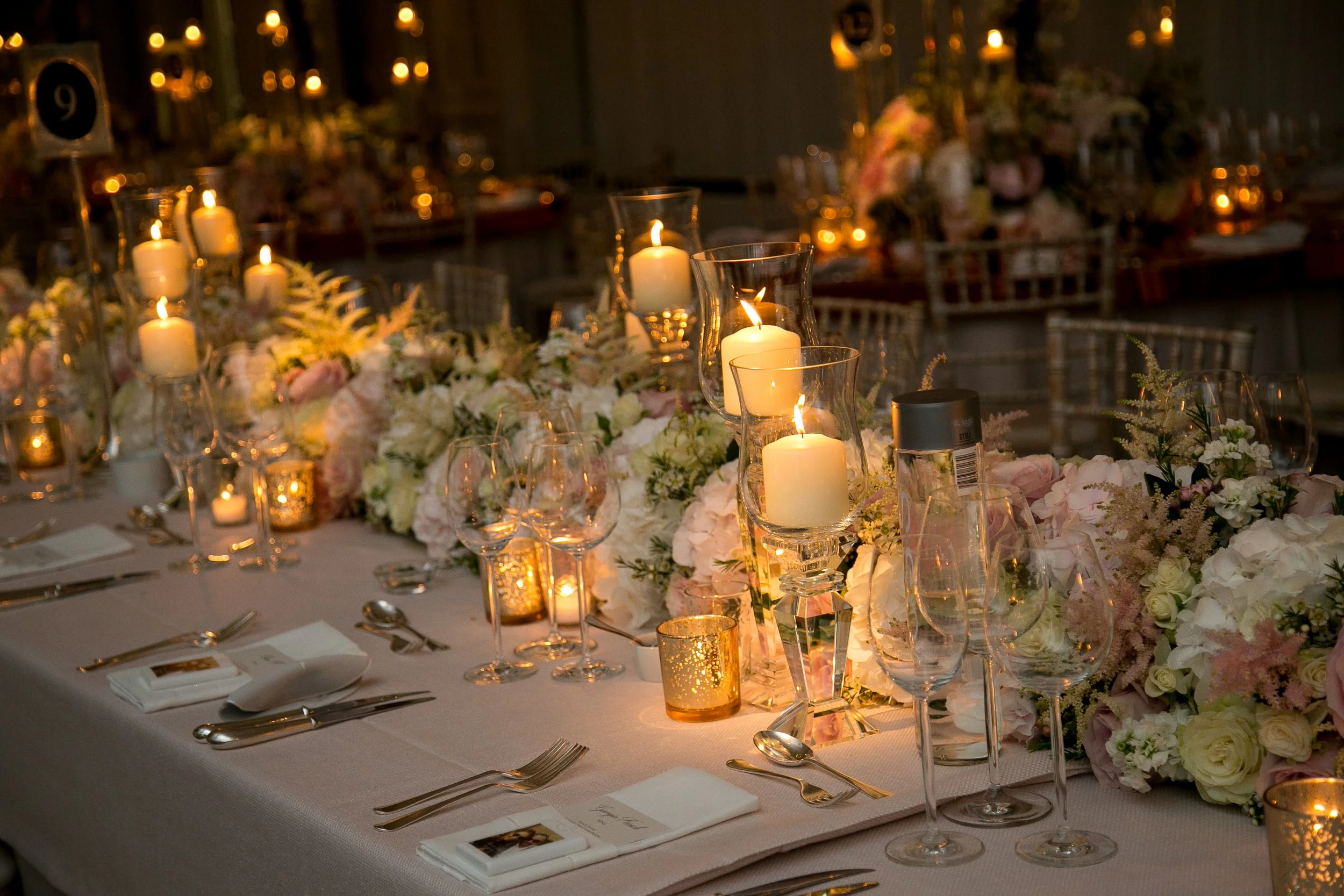 Elegant wedding reception table decorated with white flowers, lit candles in glass holders, glassware, and silverware in a dimly lit room with warm lighting.