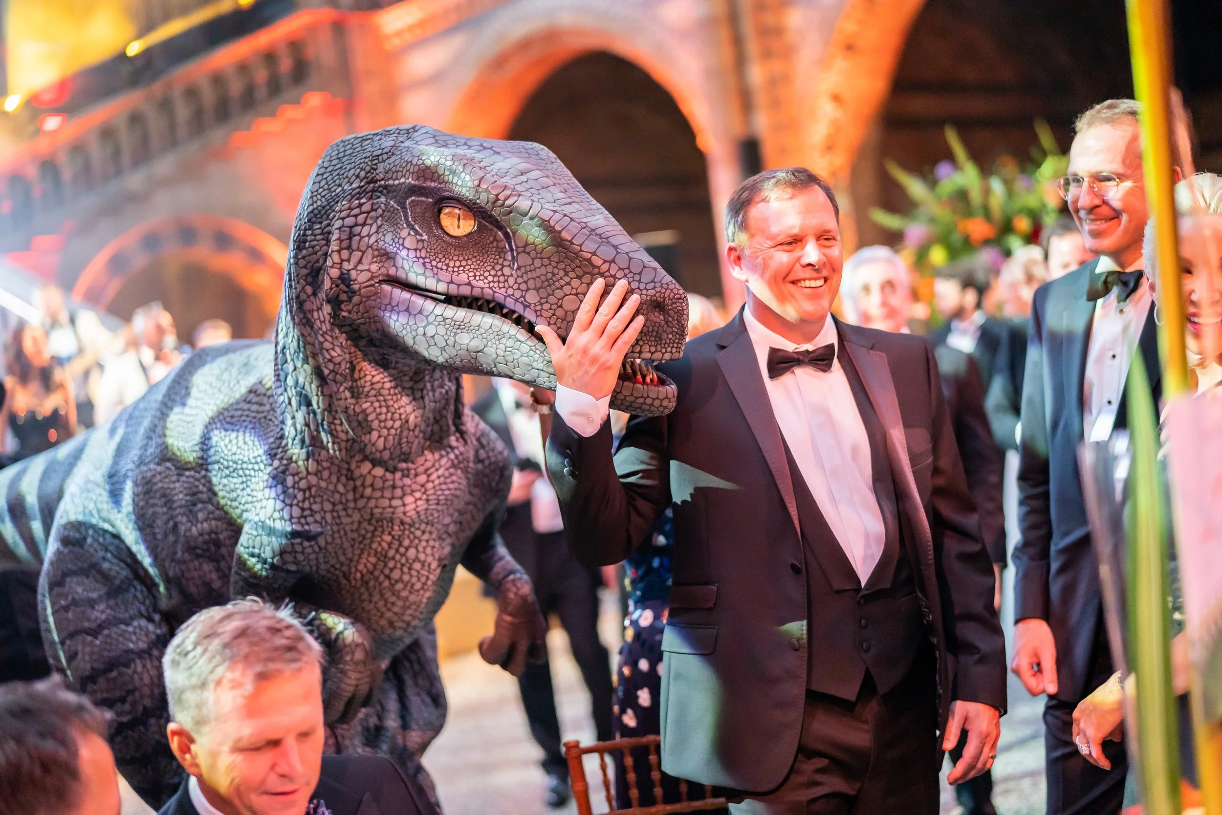 People dressed in formal attire, including a man in a tuxedo and bow tie and dinosaur,  at a corporate awards event with a decorated background and some guests in the background at The Natural History Museum.