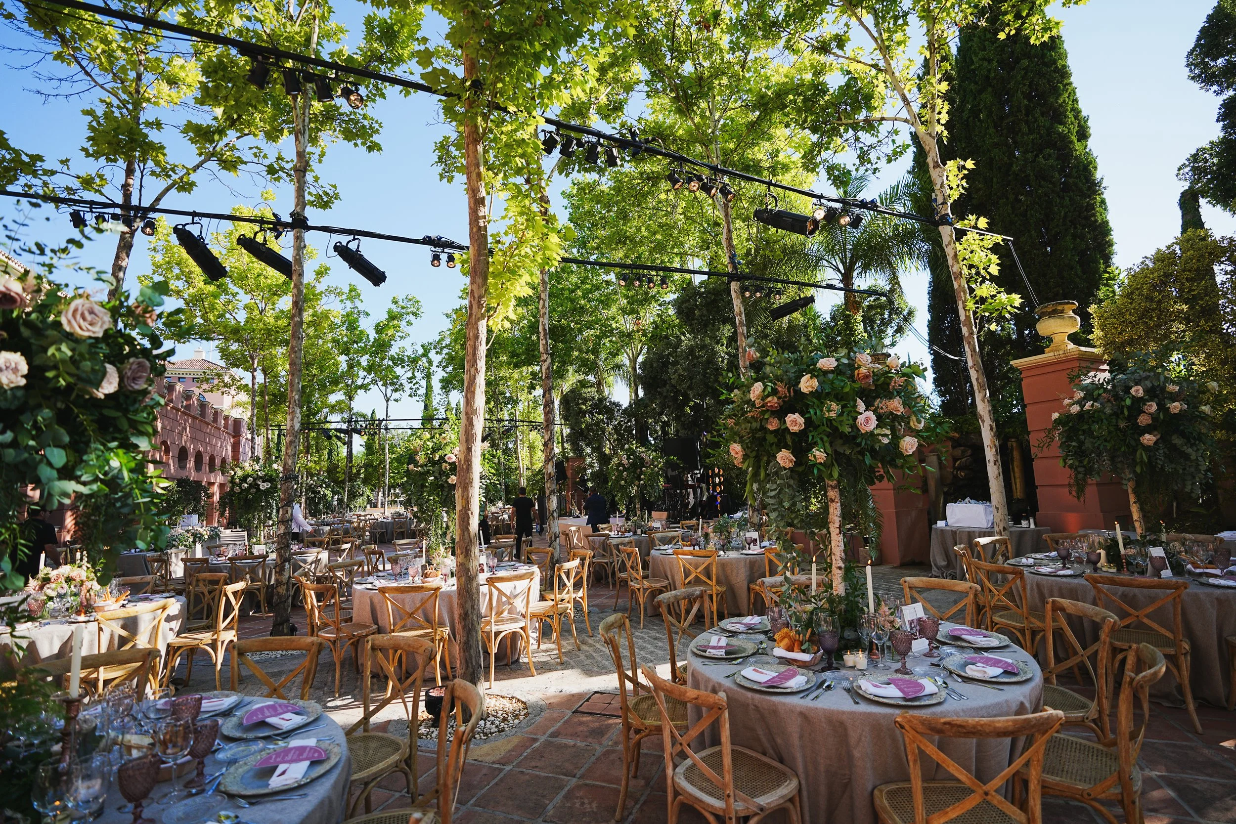 Outdoor event setup with round tables covered with white tablecloths, decorated with purple, orange, and pink details, surrounded by wooden chairs, under a canopy of green trees with string lights overhead, in a sunny setting with a pink wall and lar