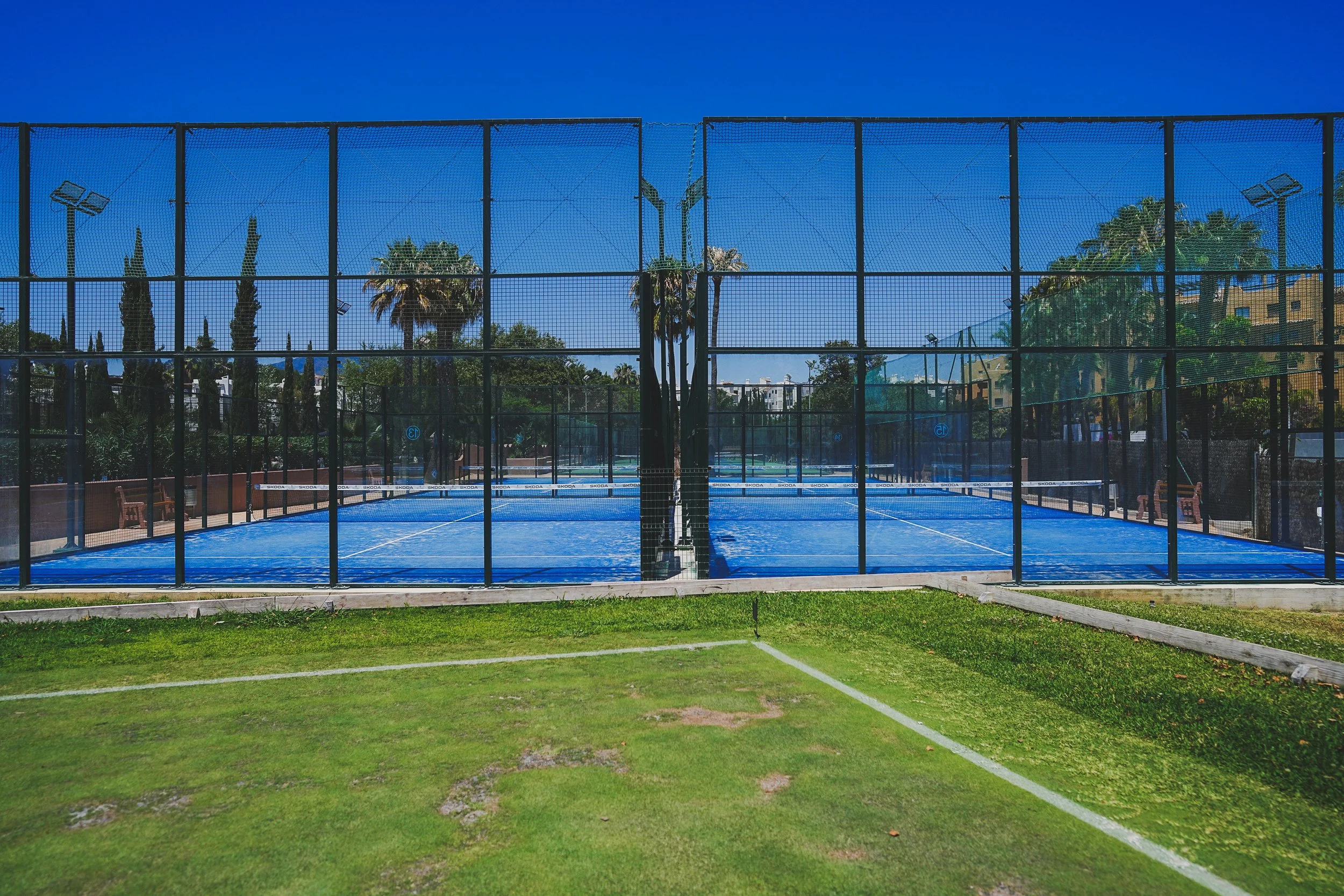 An outdoor tennis court enclosed by a black mesh fence, with palm trees, other greenery, and a building with several windows in the background, under a clear blue sky.