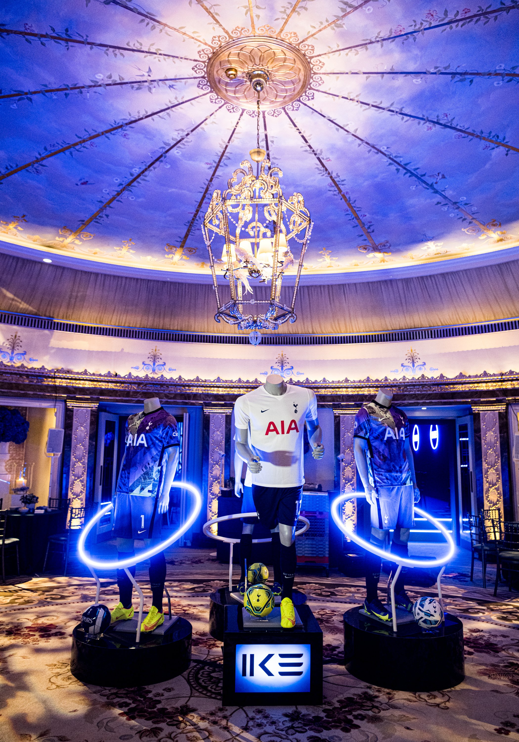 Display of Tottenham Hotspur football jerseys and equipment in a luxurious room with a decorative ceiling and chandelier, illuminated by blue and warm lighting.
